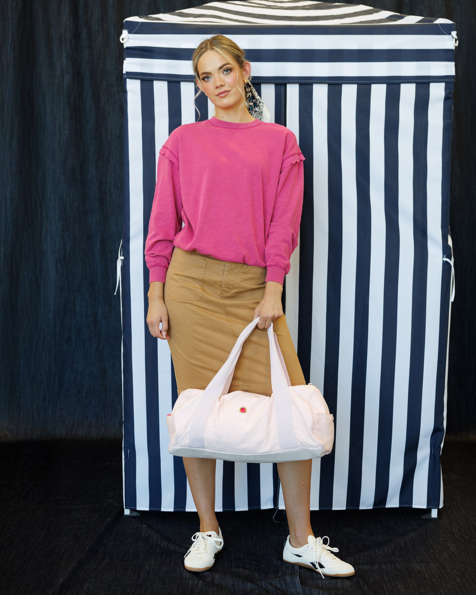 A woman stands in front of a black and white vertically striped backdrop, wearing a bright pink long-sleeve top, a tan pencil skirt, and white sneakers, while holding a large pale pink duffel bag with white handles.