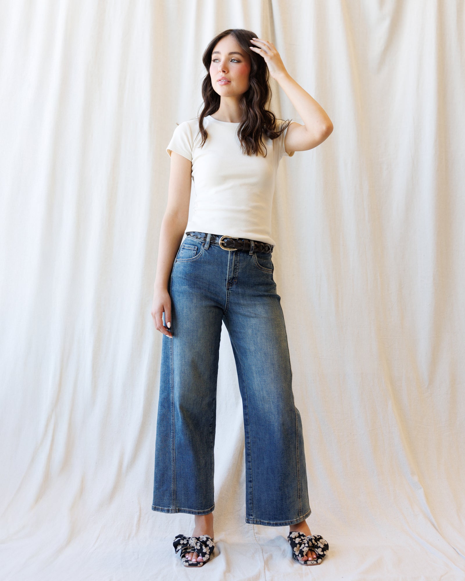 A woman modeling a pair of wide-leg blue jeans with a black belt, paired with a cream t-shirt and floral flat shoes, standing against a draped white fabric backdrop.