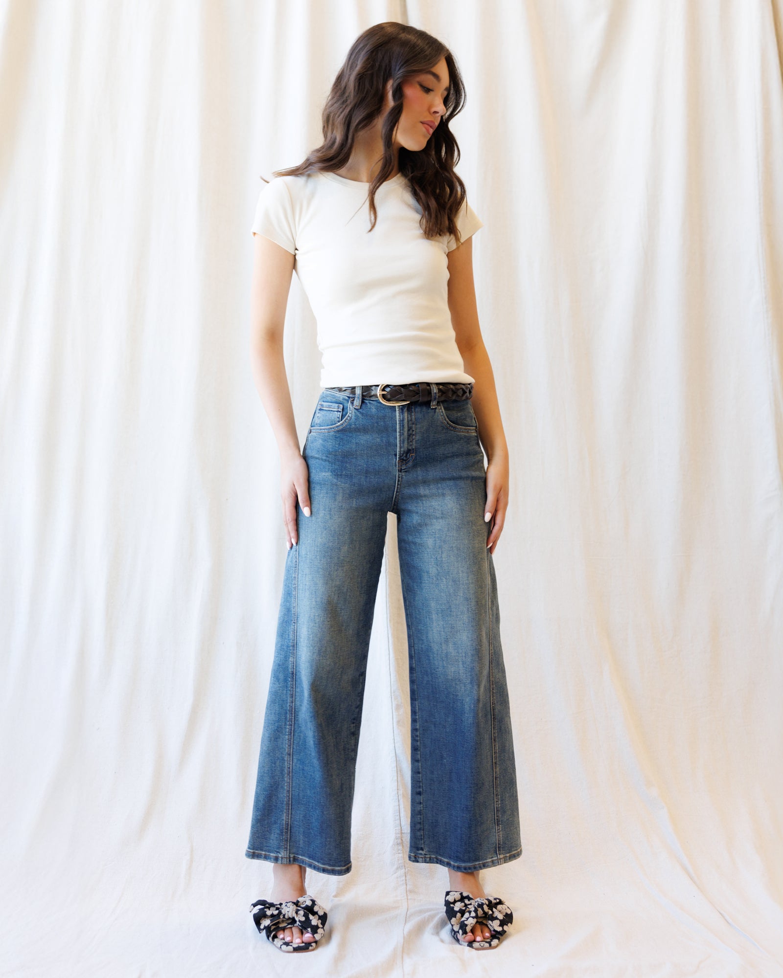 A woman modeling a pair of wide-leg blue denim jeans with a black belt, styled with a white t-shirt and floral print slippers against a draped white fabric backdrop.