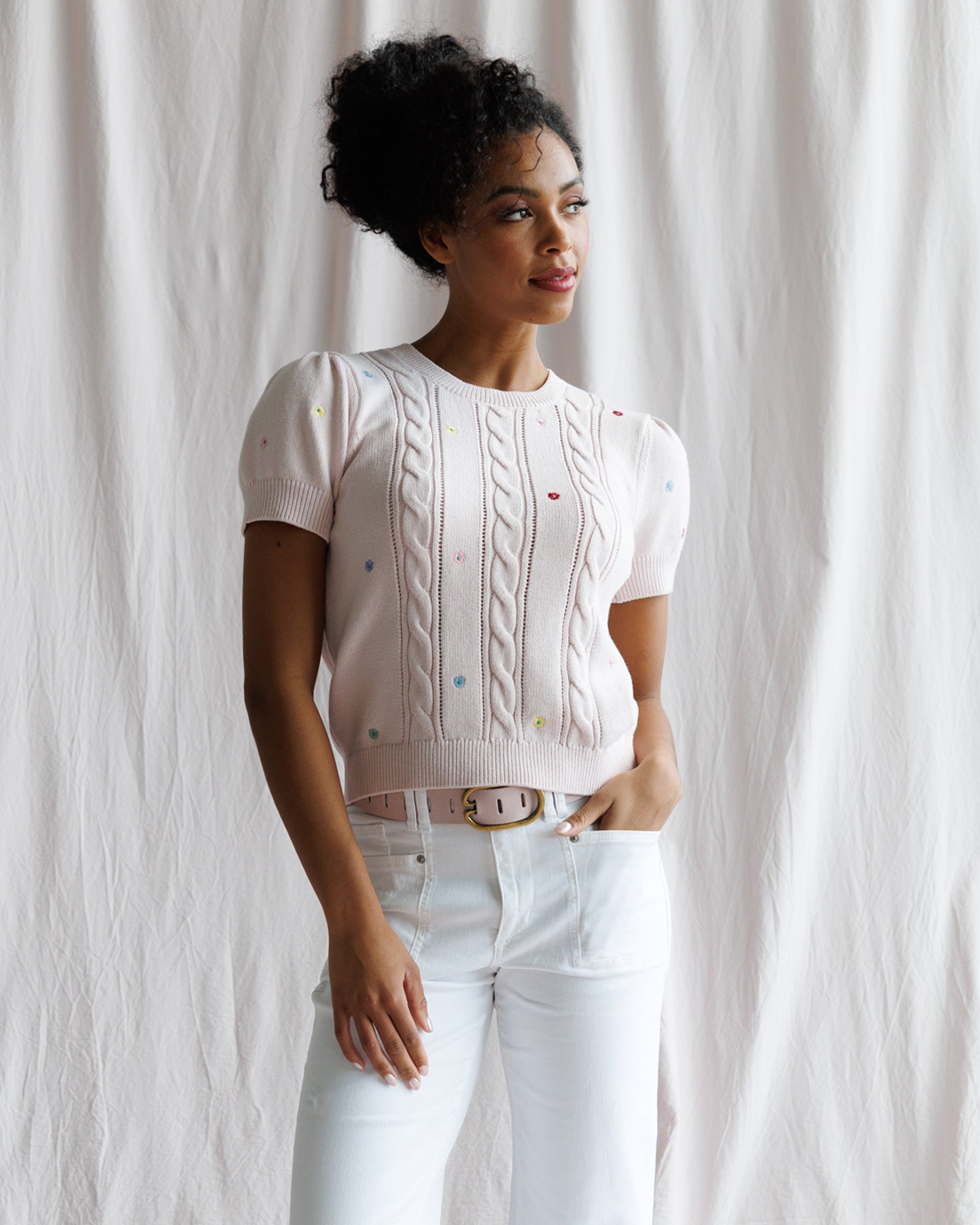 Woman wearing a pink cable knit sweater and white pants against a white curtain background