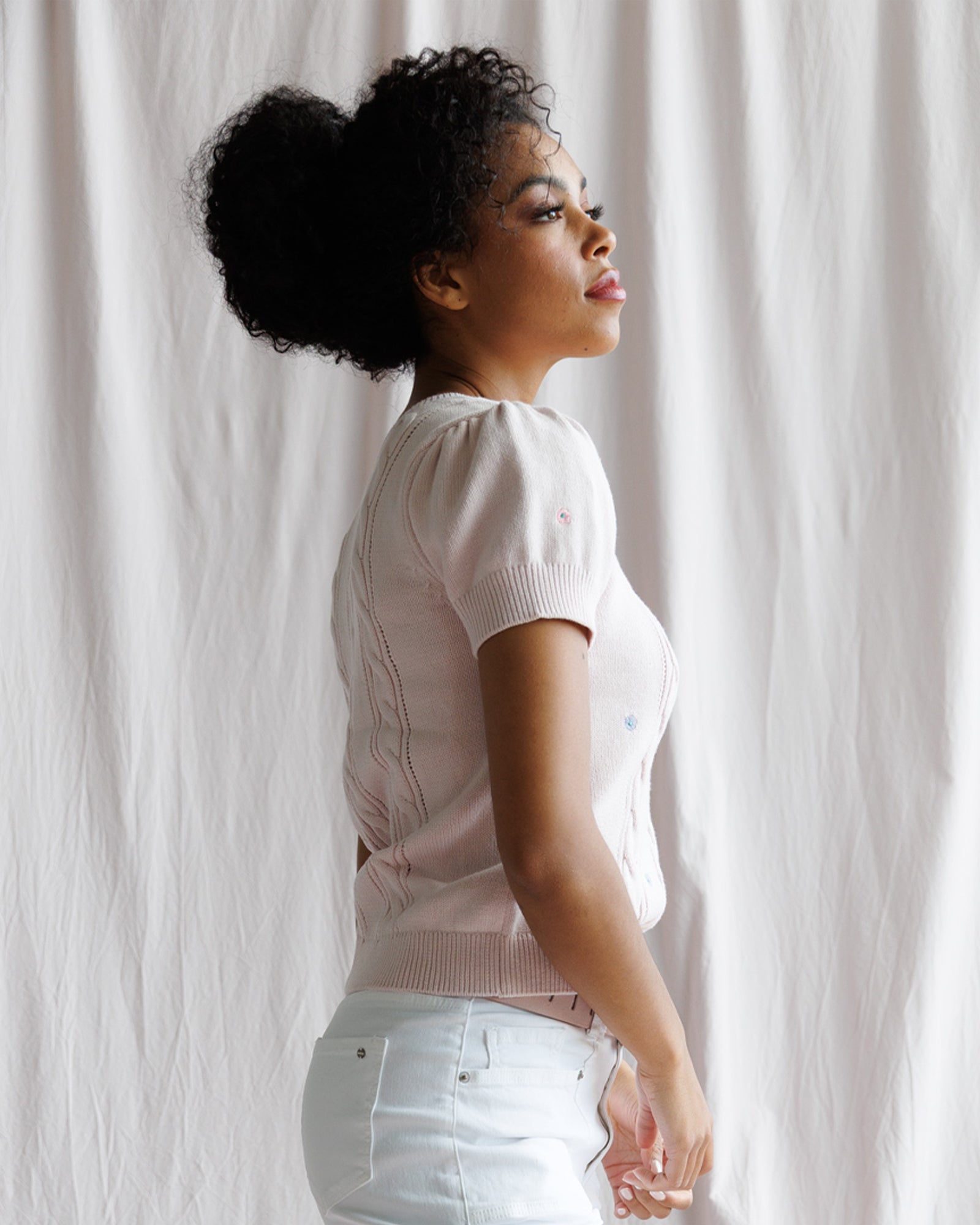 Woman in a light pink sweater and white pants standing against a white curtain.