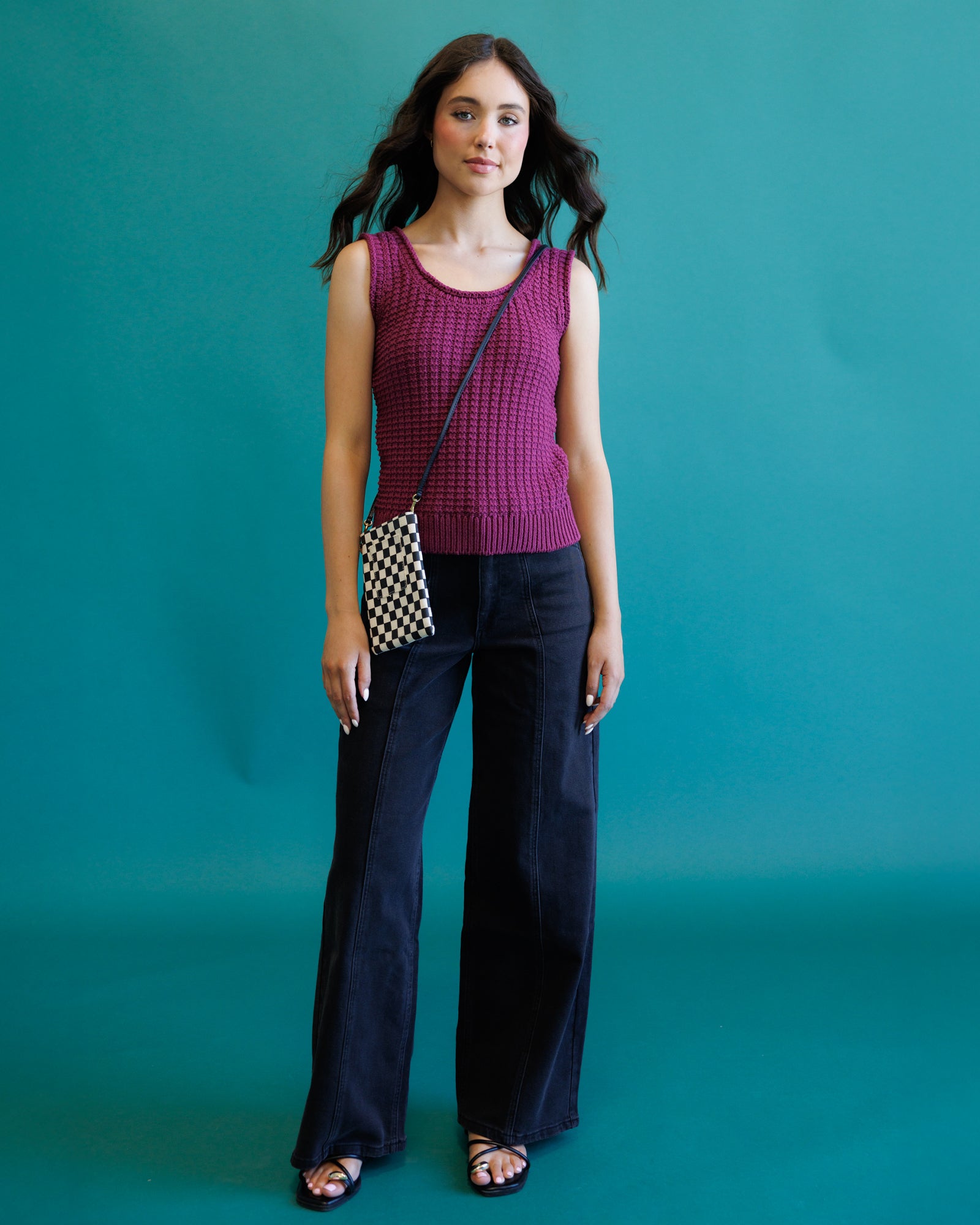 A woman stands against a solid teal background wearing a sleeveless, textured magenta top, dark wide-leg jeans, and black sandals, while carrying a small black and white checkered crossbody bag.