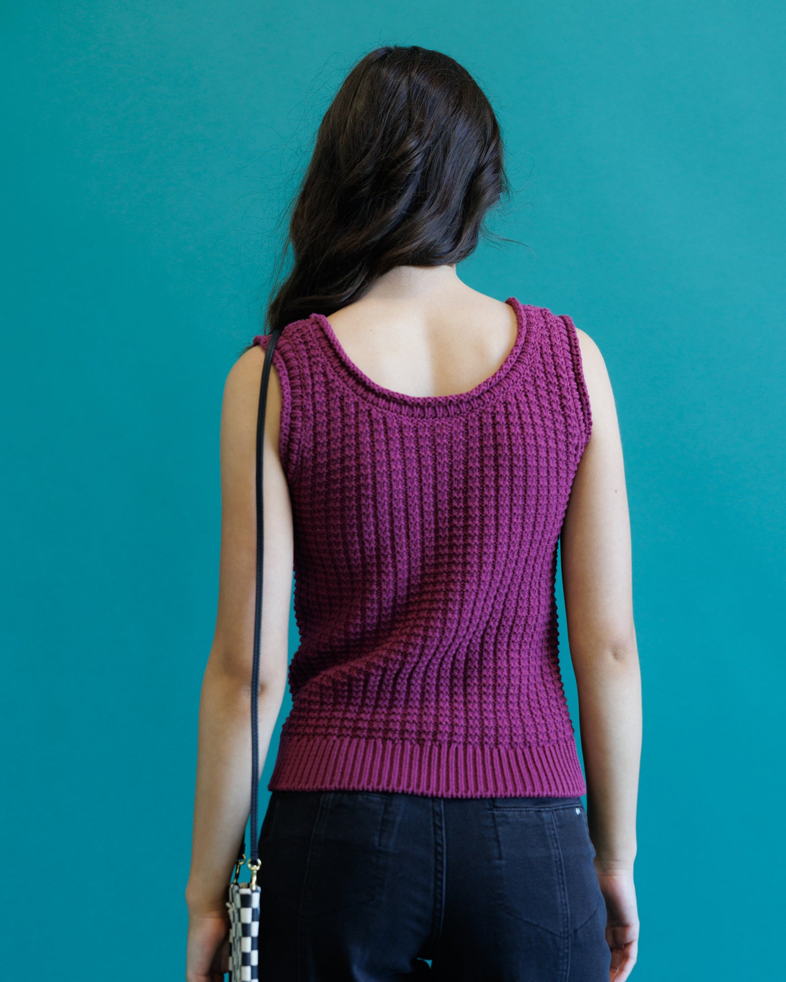 A woman shown from behind wearing a sleeveless, textured knit top in a deep magenta-purple color, paired with dark jeans, set against a solid teal background.