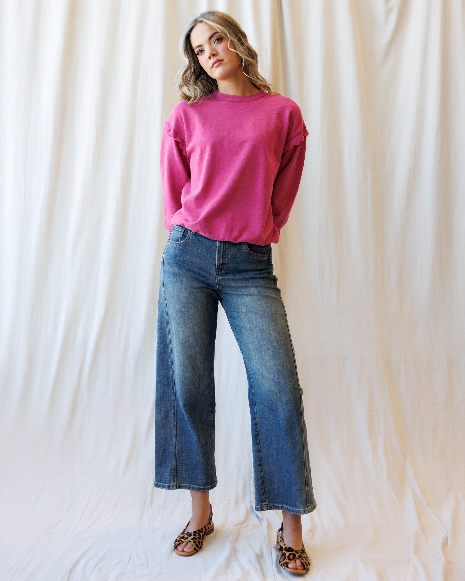 A woman modeling a bright pink long-sleeve crewneck sweatshirt with wide-leg blue jeans and leopard-print sandals, standing against a draped white fabric backdrop.