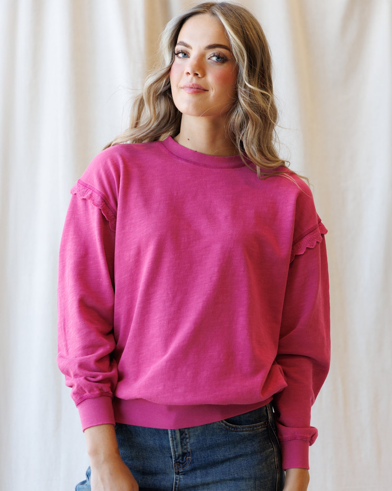 A woman wearing a bright pink crewneck sweatshirt with ruffle details on the shoulders and cuffs, paired with blue jeans, standing against a soft beige fabric backdrop.
