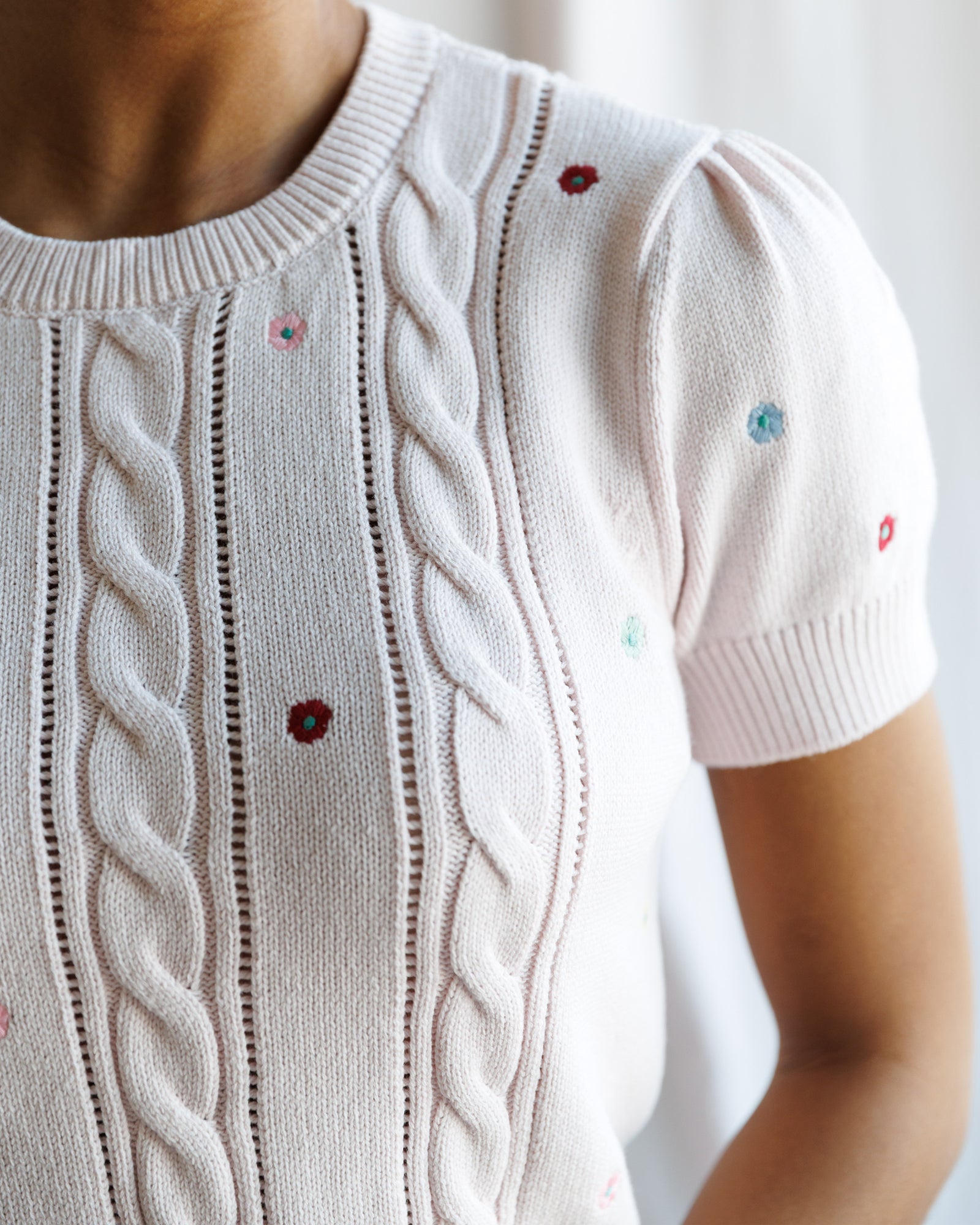 Close-up of a white knit sweater with cable patterns and embroidered flowers.
