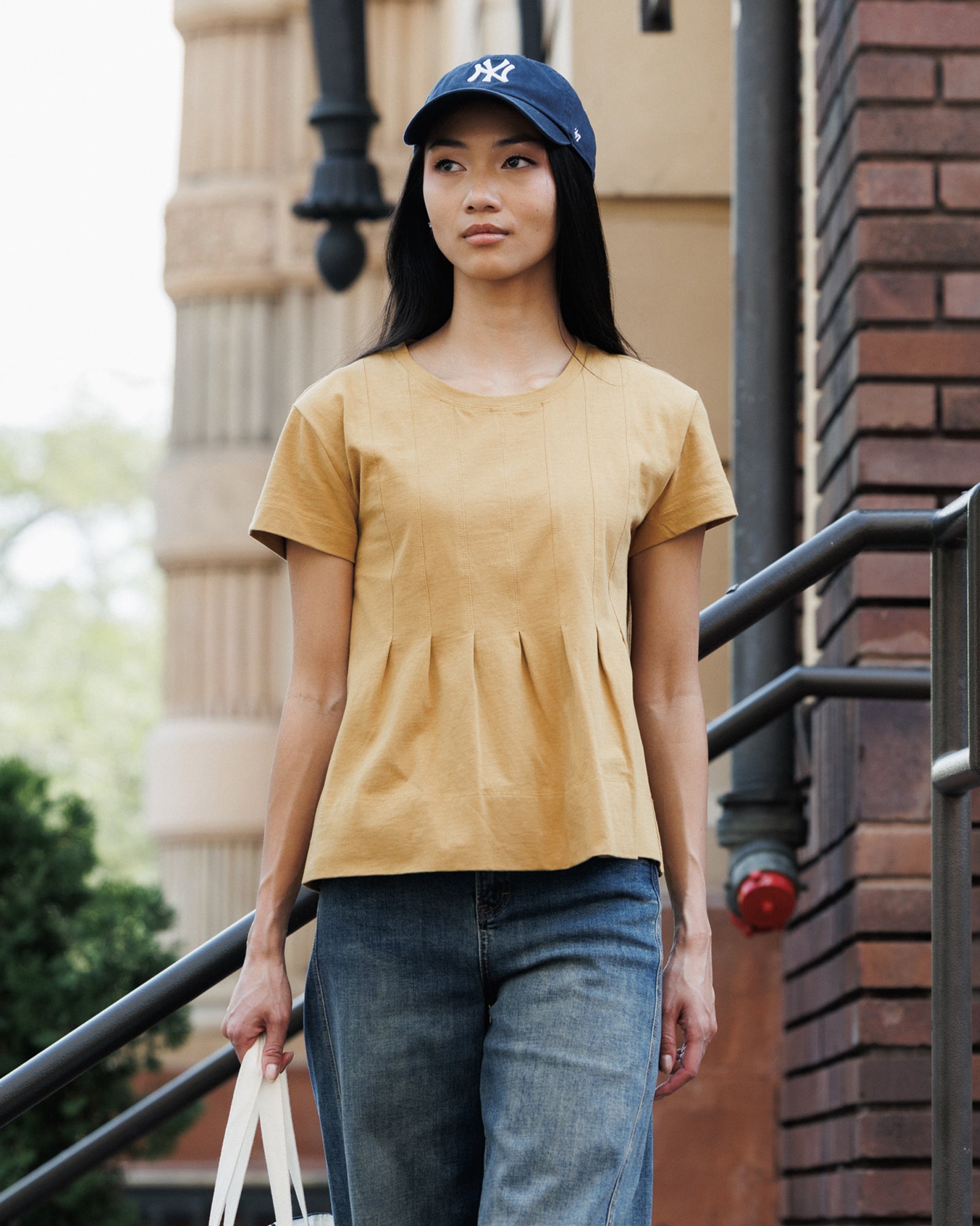 A woman wearing a mustard yellow short-sleeve peplum top, blue jeans, and a navy blue New York Yankees baseball cap, standing on outdoor stairs.
