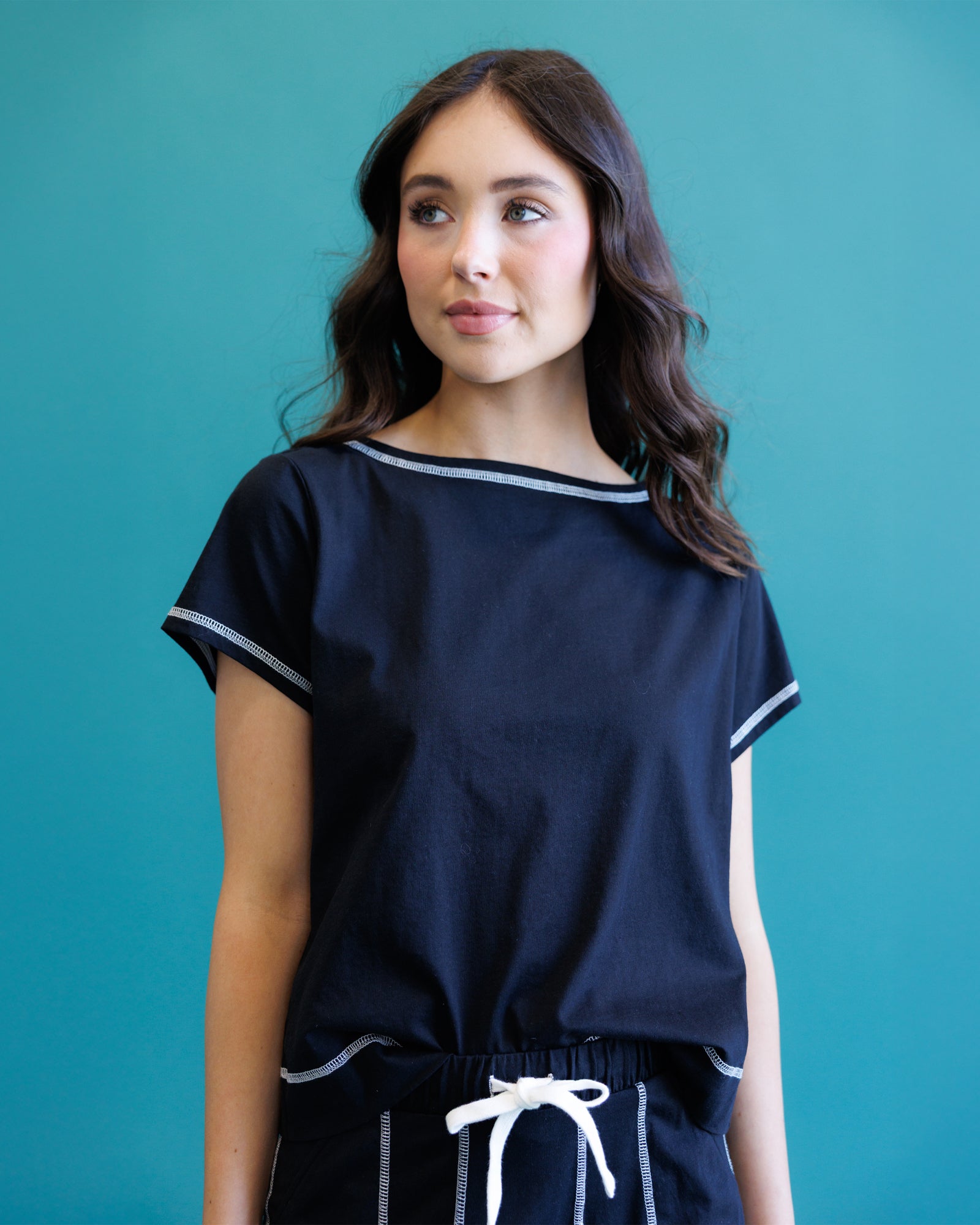 A woman modeling a black short-sleeve top with white contrast trim and a wide boat neckline, paired with matching dark bottoms, set against a solid teal background.