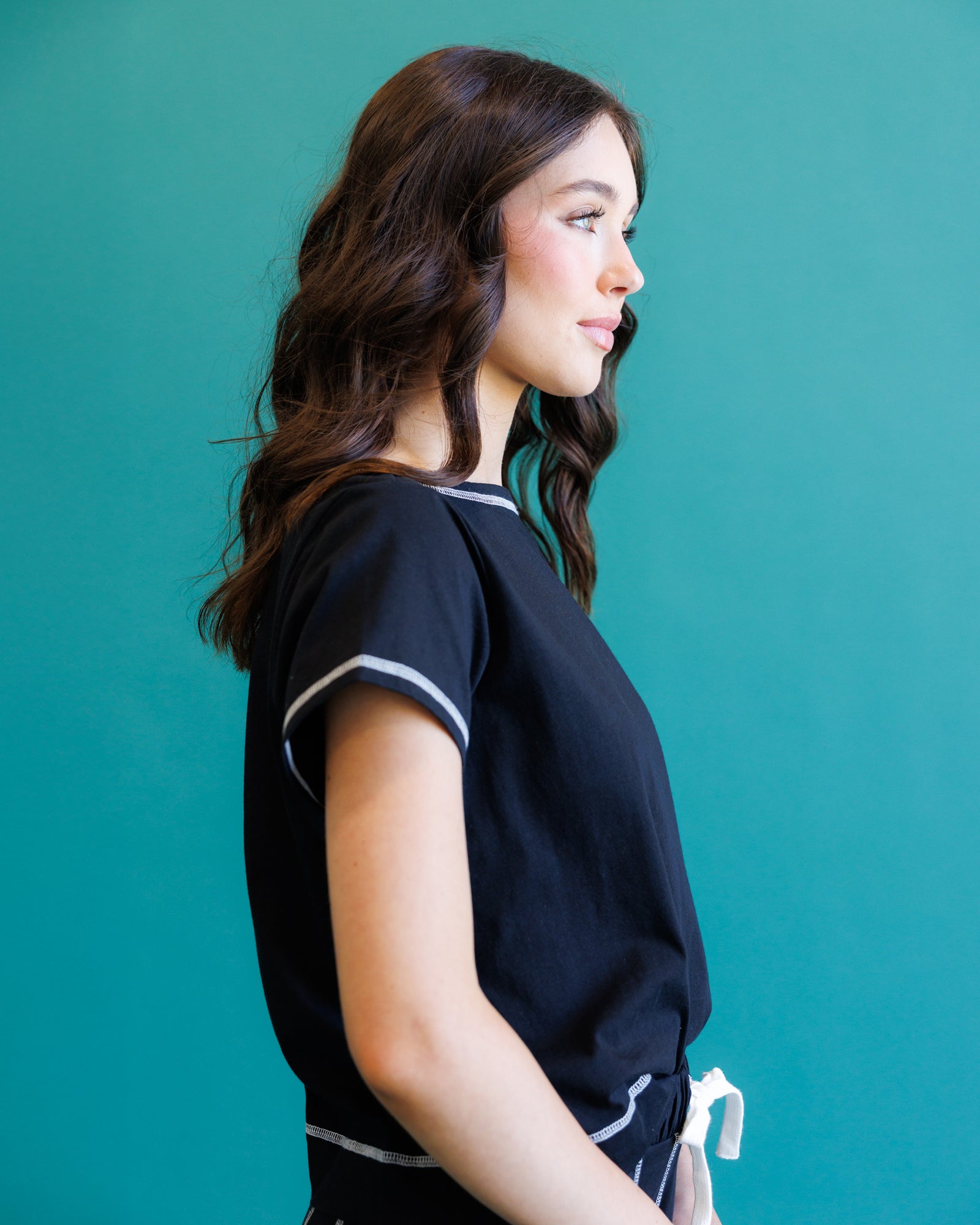 A woman in profile wearing a black short-sleeve top with white contrast trim, standing against a solid teal background.