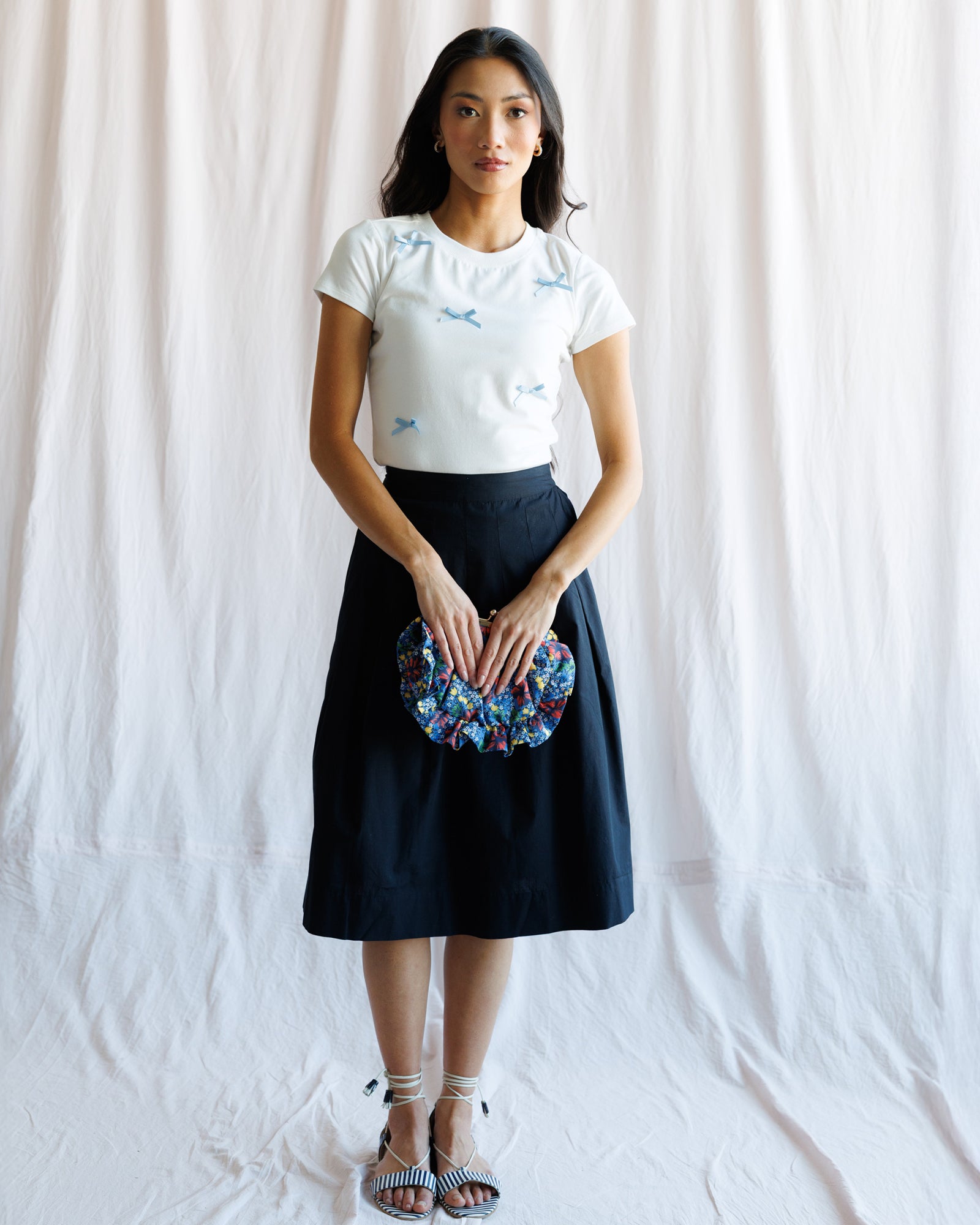 Woman wearing a white t-shirt with blue graphics and a navy skirt, holding a colorful clutch against a plain background.