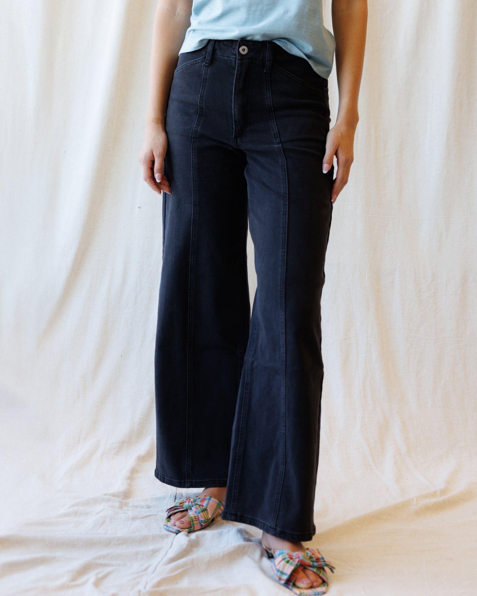 A person wearing black wide-leg denim pants with a light blue top and patterned flat sandals, standing against a draped cream fabric backdrop.