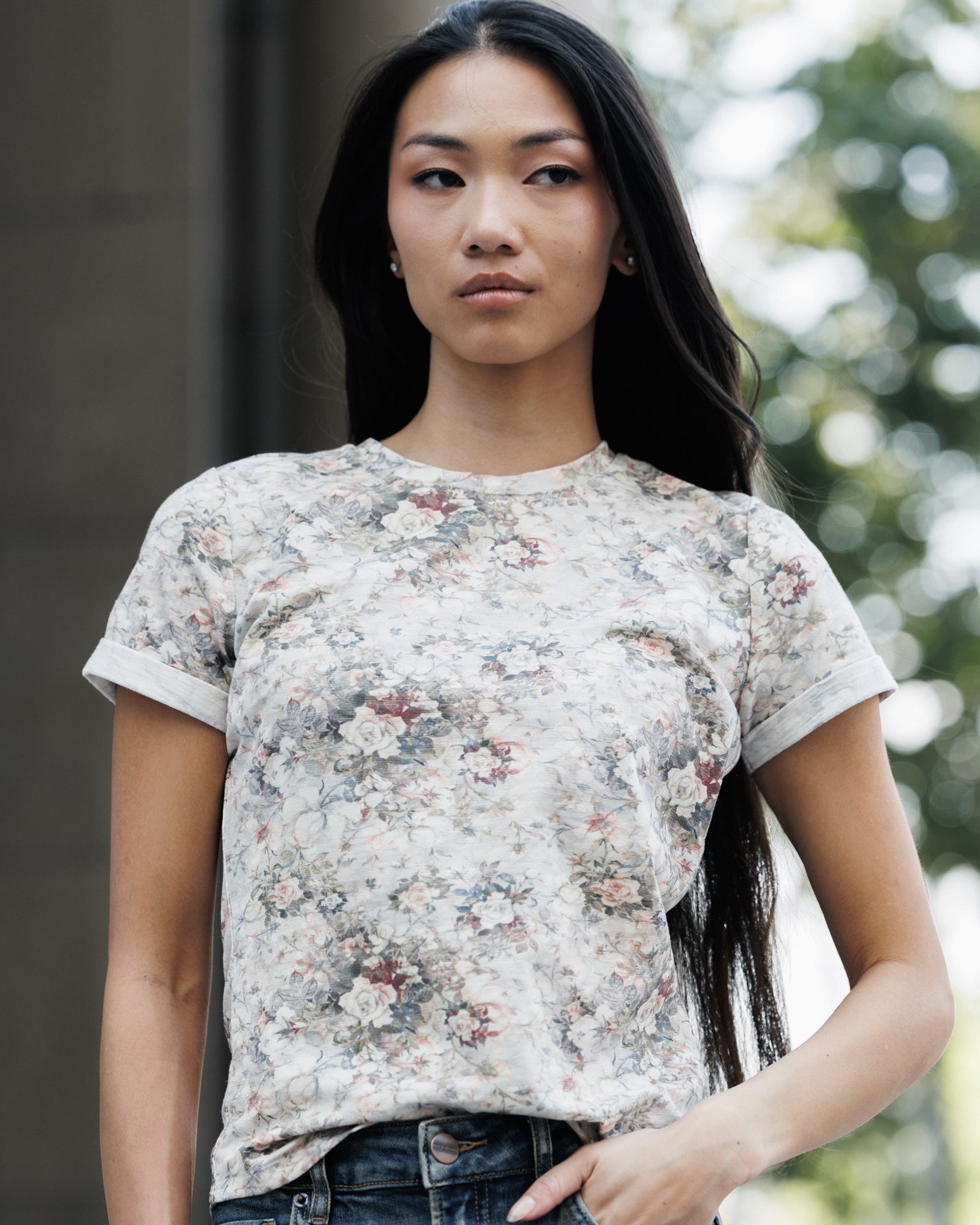 A woman wearing a short-sleeve crewneck t-shirt with a faded vintage floral pattern in muted pink, blue, and green tones on a light beige background, paired with dark blue jeans.