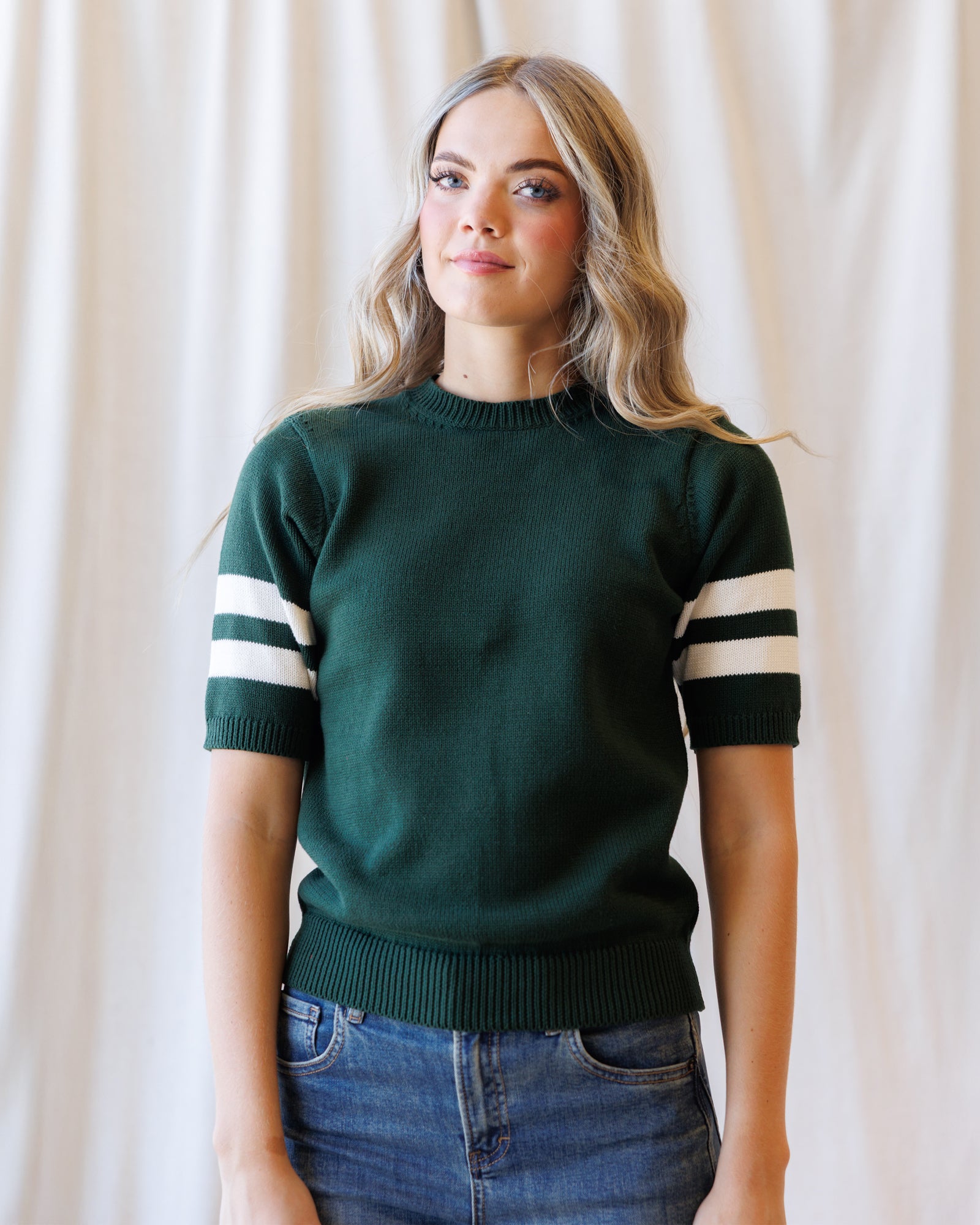 A woman wearing a dark green short-sleeve knit sweater with white stripes on the sleeves, paired with blue jeans, standing against a draped cream fabric backdrop.
