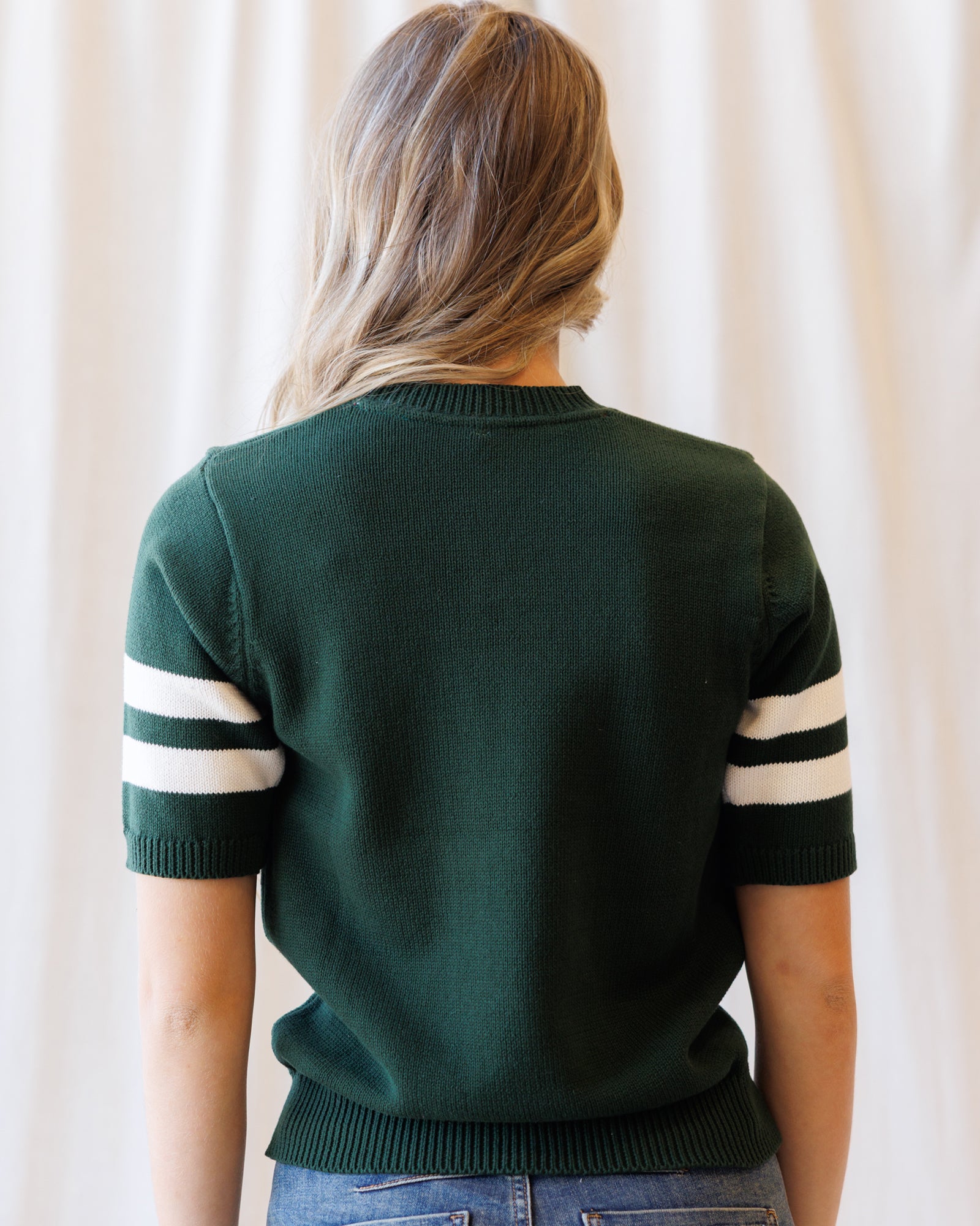 A woman viewed from behind wearing a dark green short-sleeve knit sweater with white stripes on the sleeves, paired with blue jeans against a soft cream curtain background.