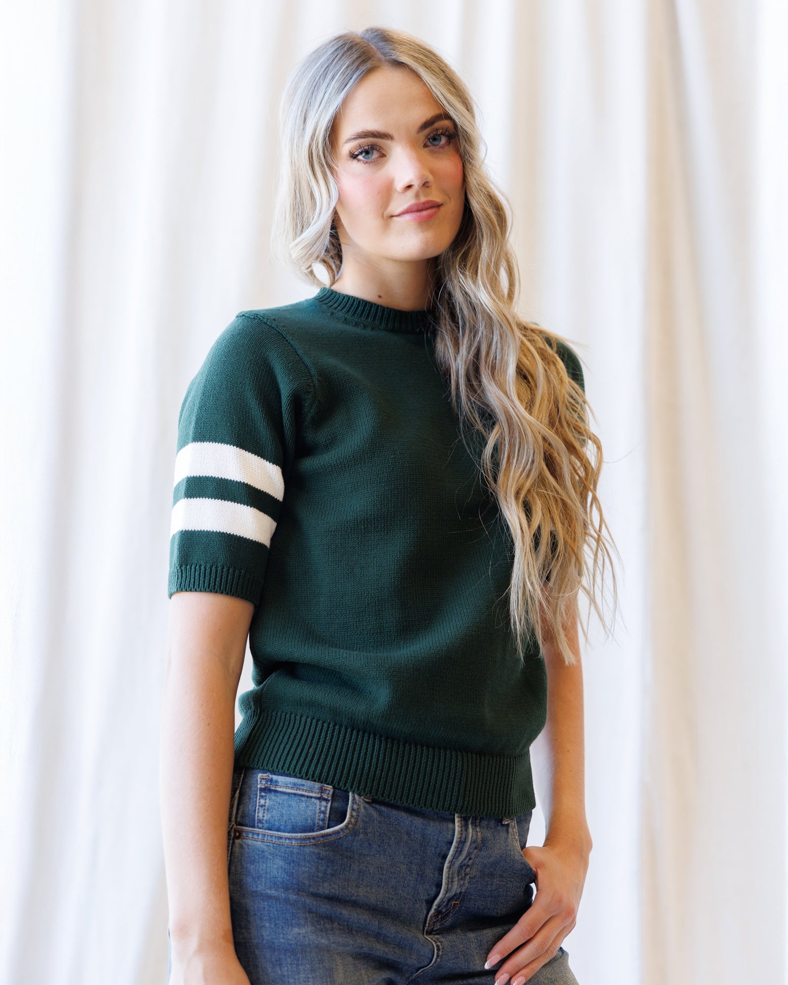 A woman with long blonde wavy hair wearing a dark green short-sleeve knit sweater with two white stripes on the sleeve, paired with blue jeans, standing in front of a soft white curtain background.