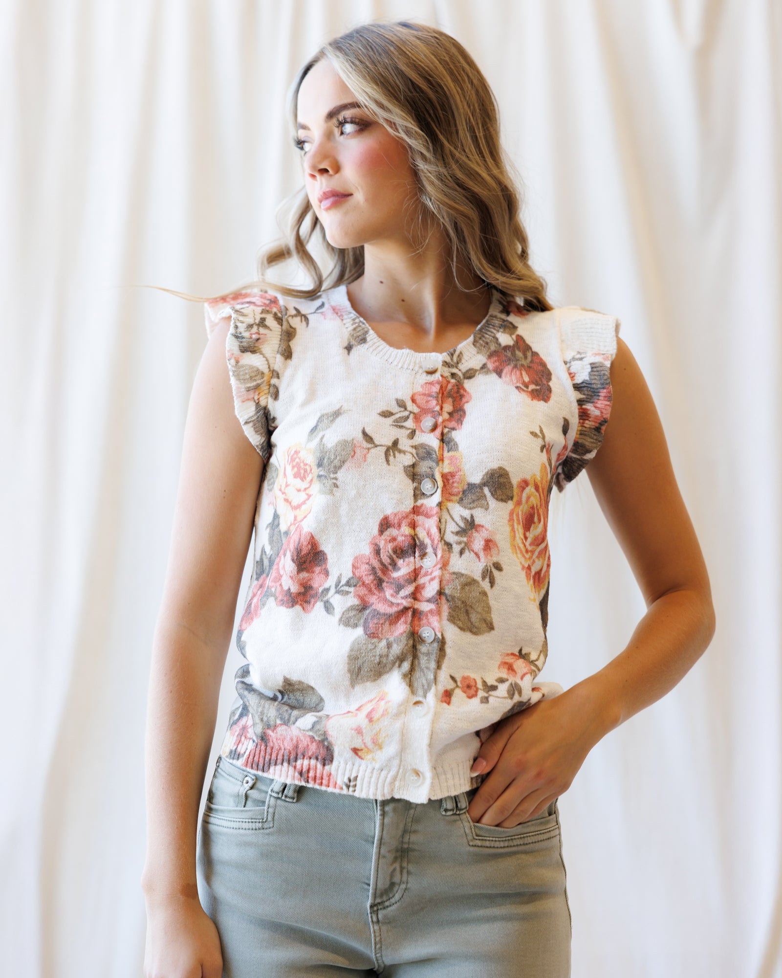 A woman wearing a sleeveless button-front top with a floral print in pink, red, and green on a cream background, paired with light gray jeans, standing against a white curtain backdrop.