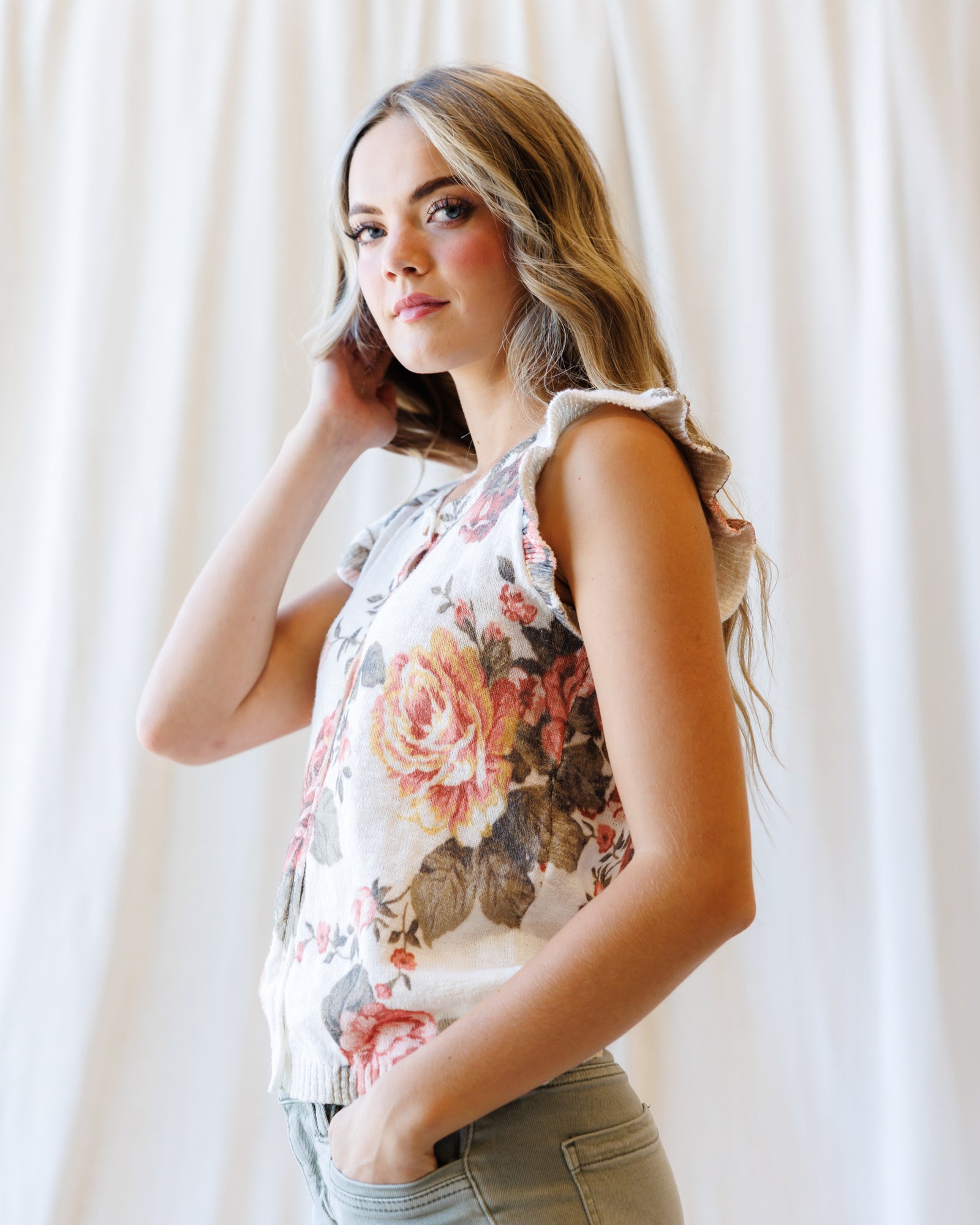 A woman wearing a sleeveless white top with a large pink and brown floral print, paired with olive green pants, standing against a white curtain background.