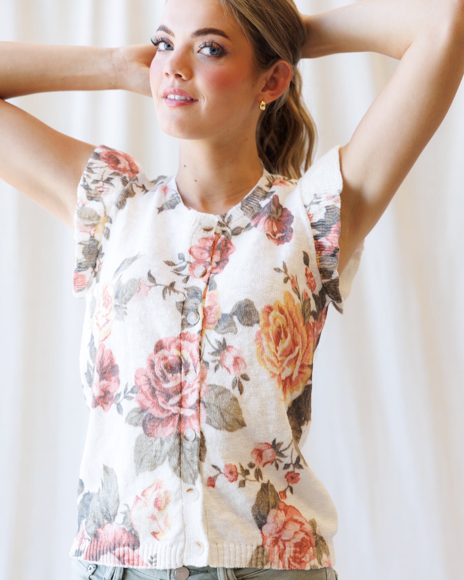A woman wearing a cream-colored sleeveless top with a pink, red, and yellow floral rose print, posing with her arms behind her head against a soft white curtain background.