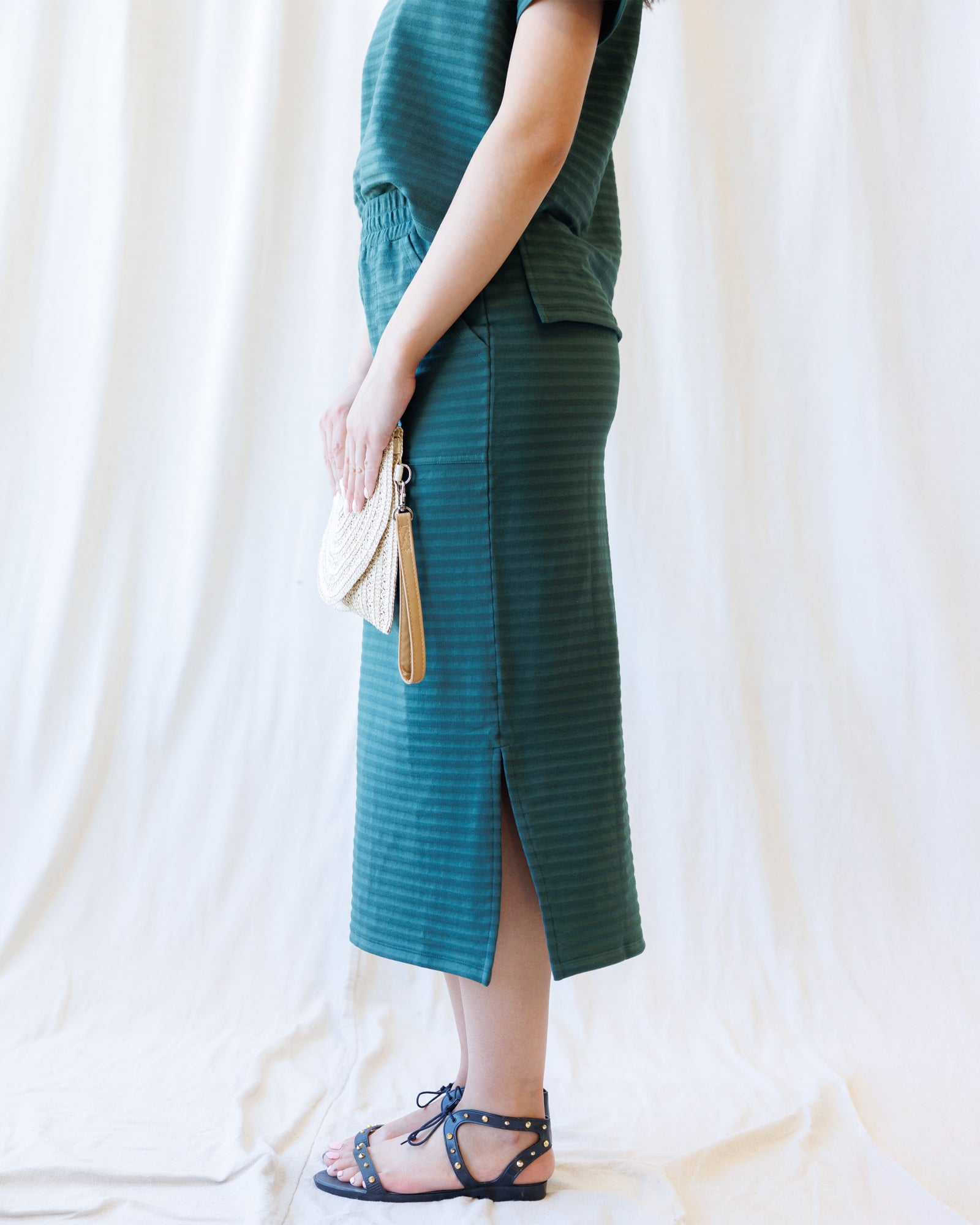 A model wearing a dark teal green striped midi skirt with a side slit, holding a woven cream clutch, and wearing black strappy sandals against a draped white fabric backdrop.
