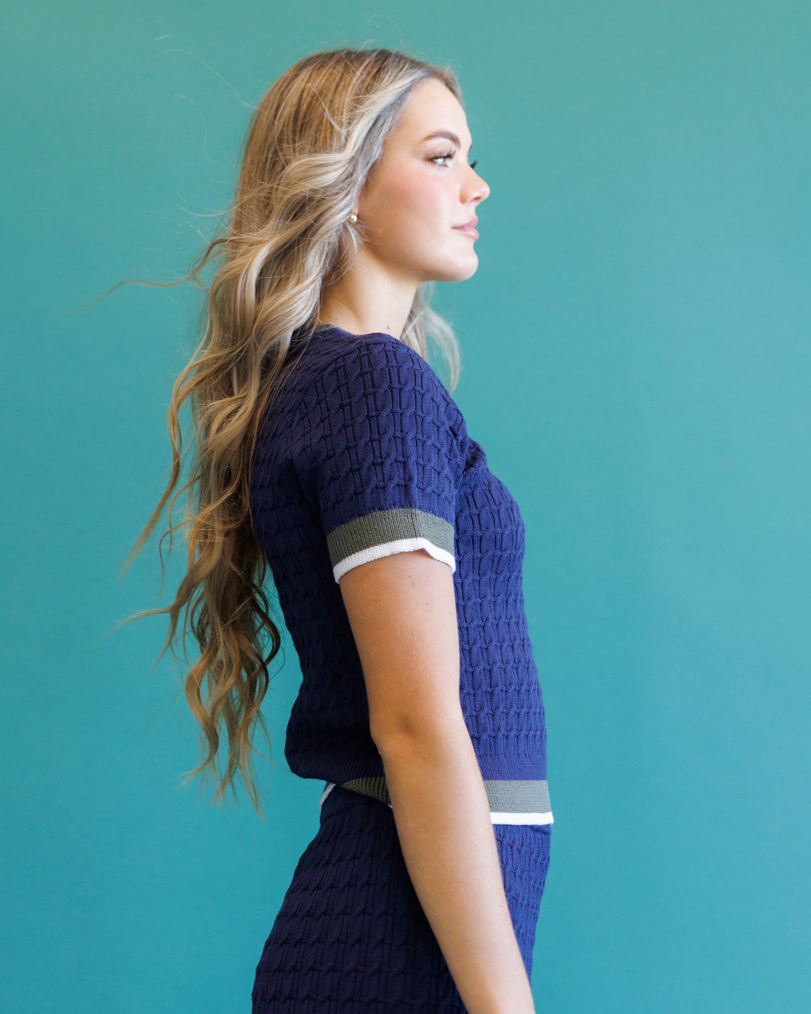 A woman in profile wearing a navy blue textured knit short-sleeve top with gray and white striped accents on the sleeve and waist, set against a solid teal background.