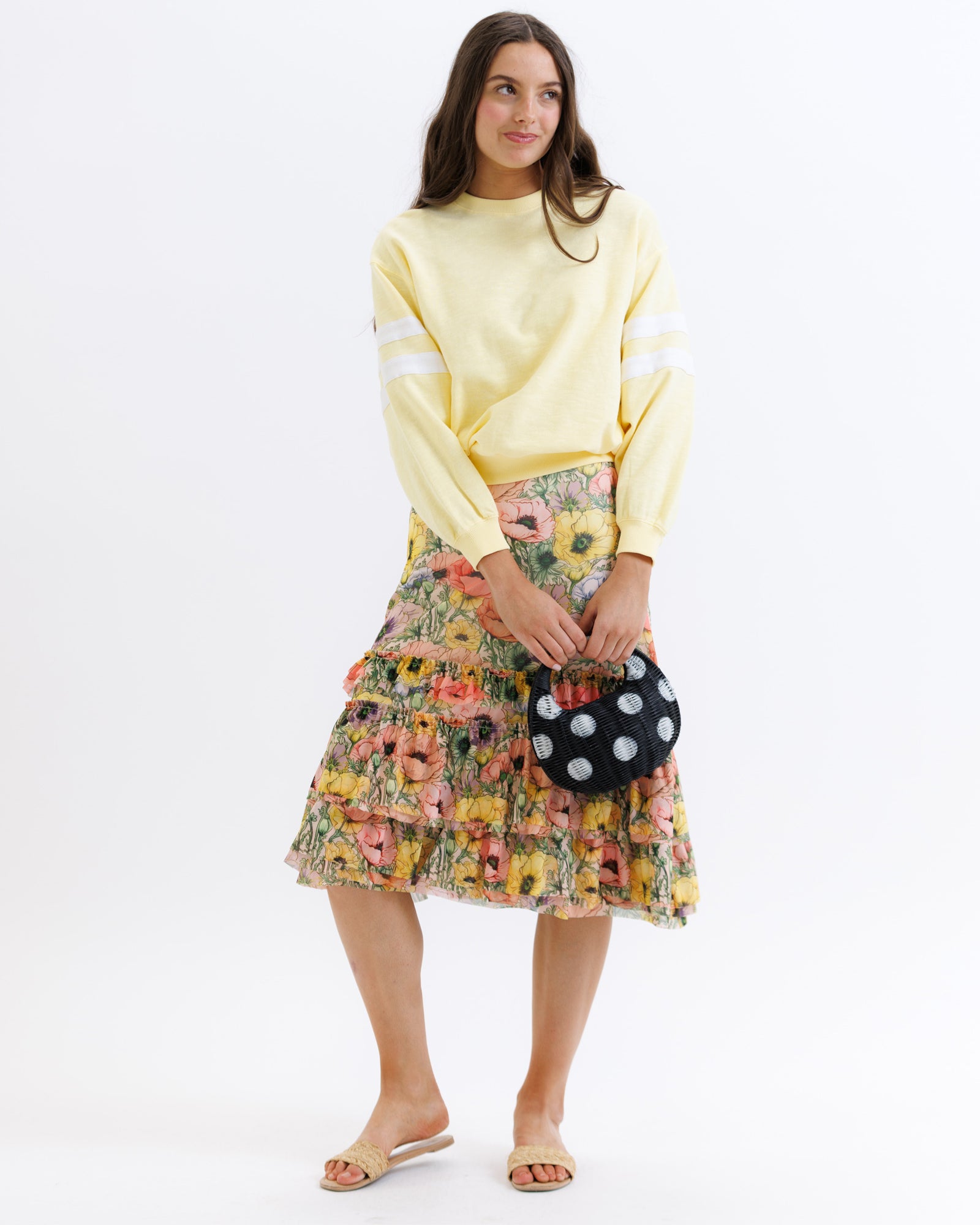 Woman wearing a yellow sweater and floral skirt holding a polka dot bag on a white background