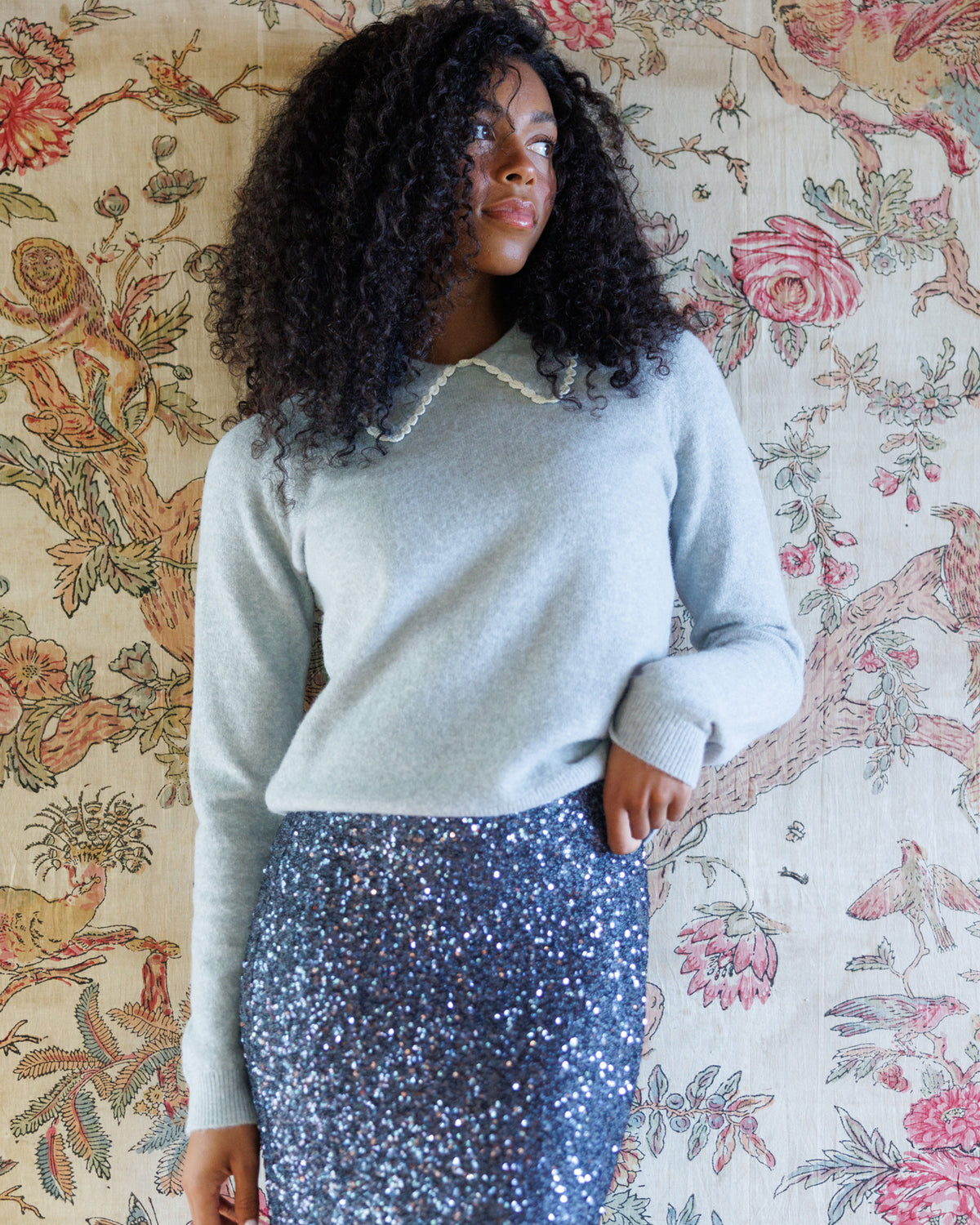 A woman with voluminous curly black hair models a light blue long-sleeve knit sweater with a white collar, paired with a sparkly blue sequined skirt, standing against a vintage floral tapestry.