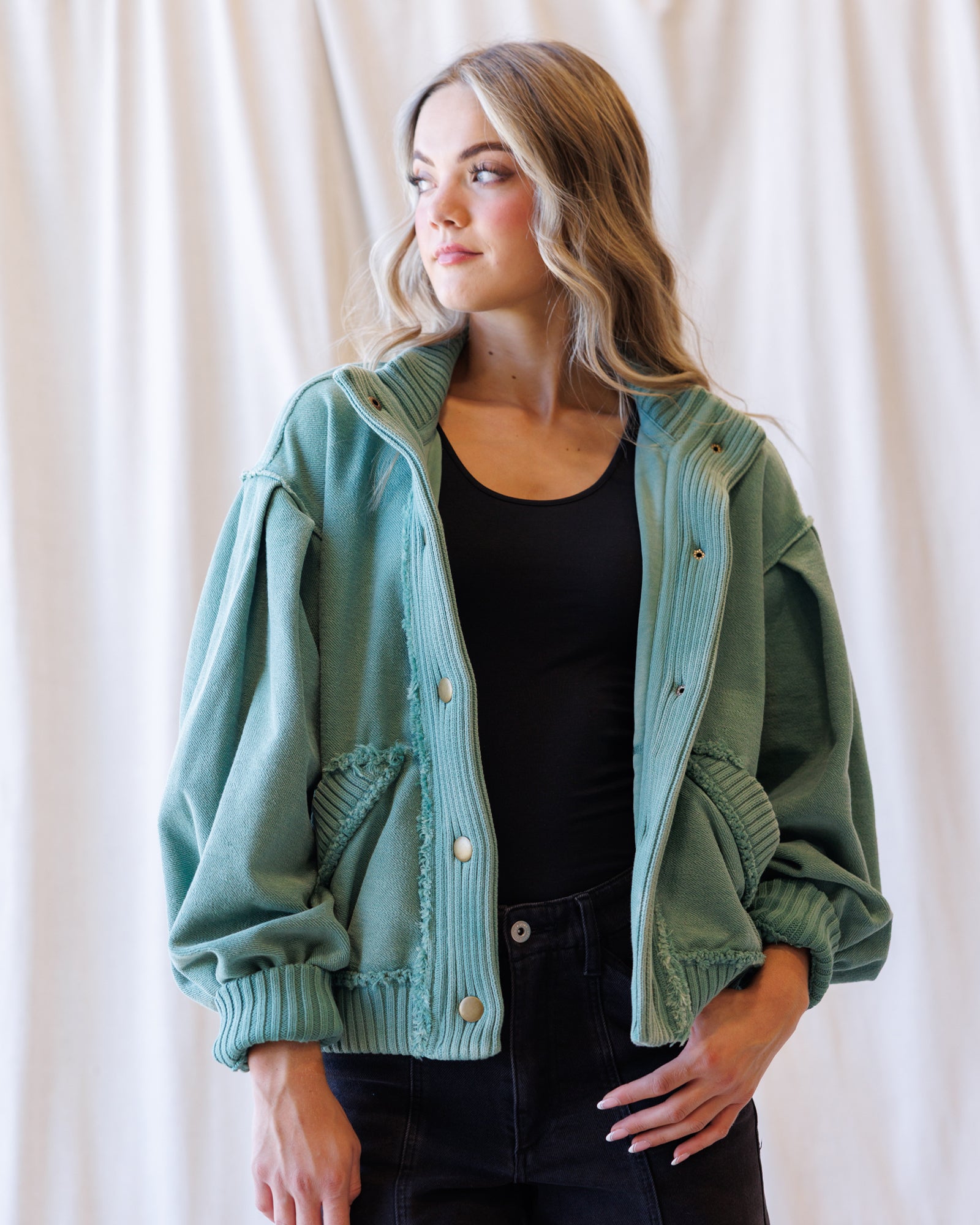 A woman wearing a sage green ribbed knit jacket with balloon sleeves and decorative buttons, layered over a black top and black jeans, standing against a draped cream fabric backdrop.