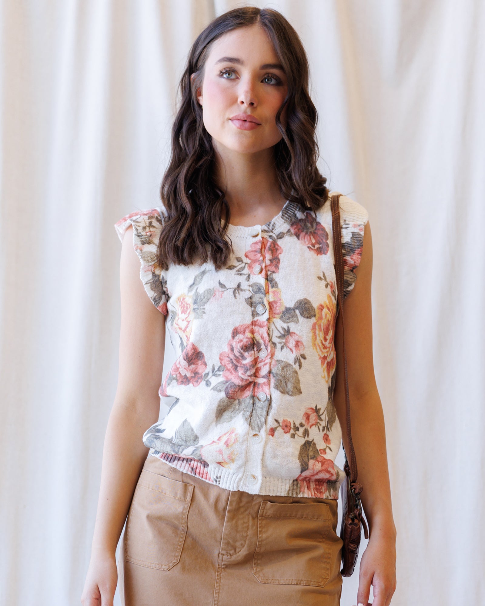 A woman wearing a sleeveless cream blouse with a pink and green floral print, paired with brown pants, standing against a soft white curtain backdrop.