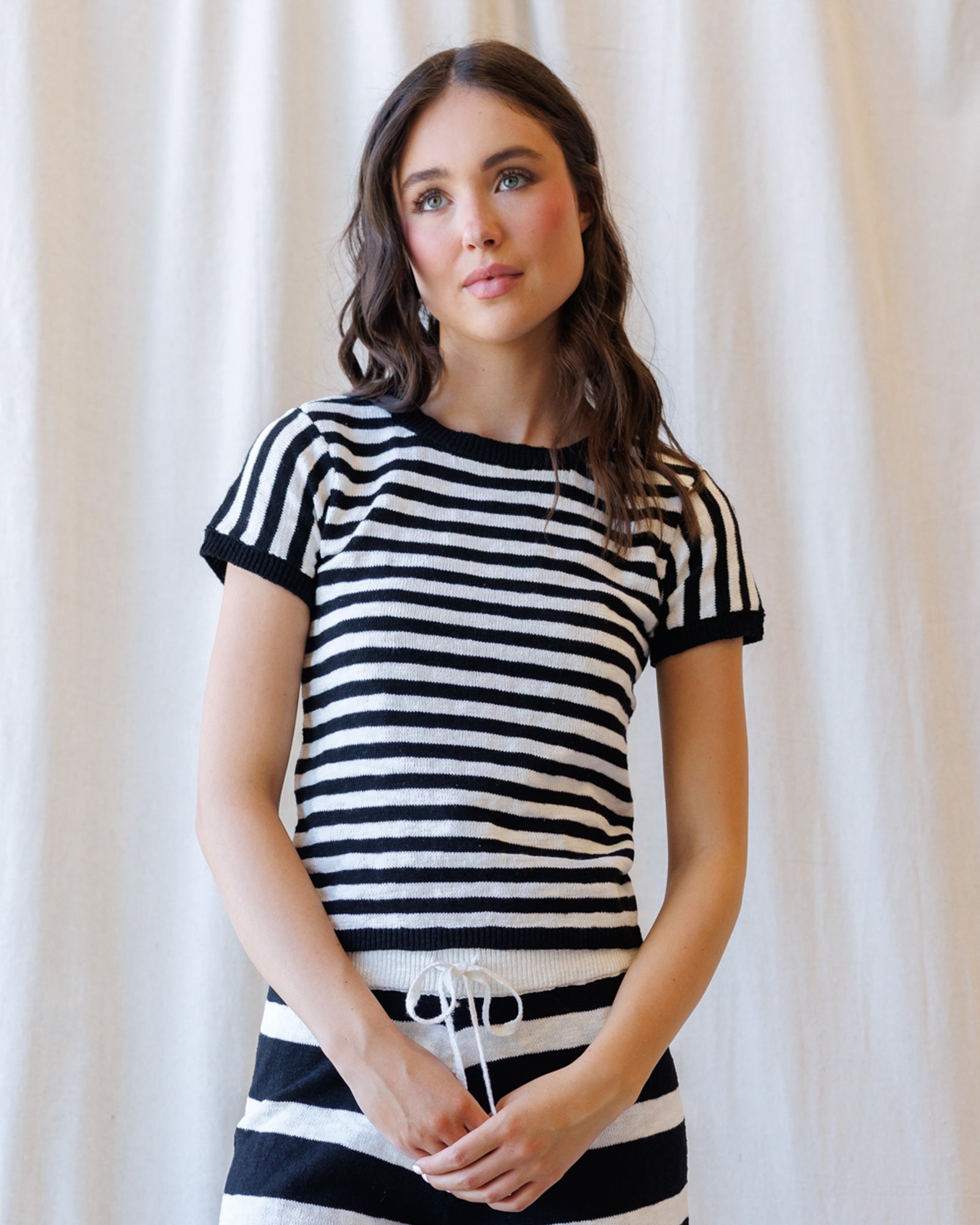 A woman wearing a short-sleeve knit top with horizontal black and white stripes, standing against a soft cream curtain backdrop.