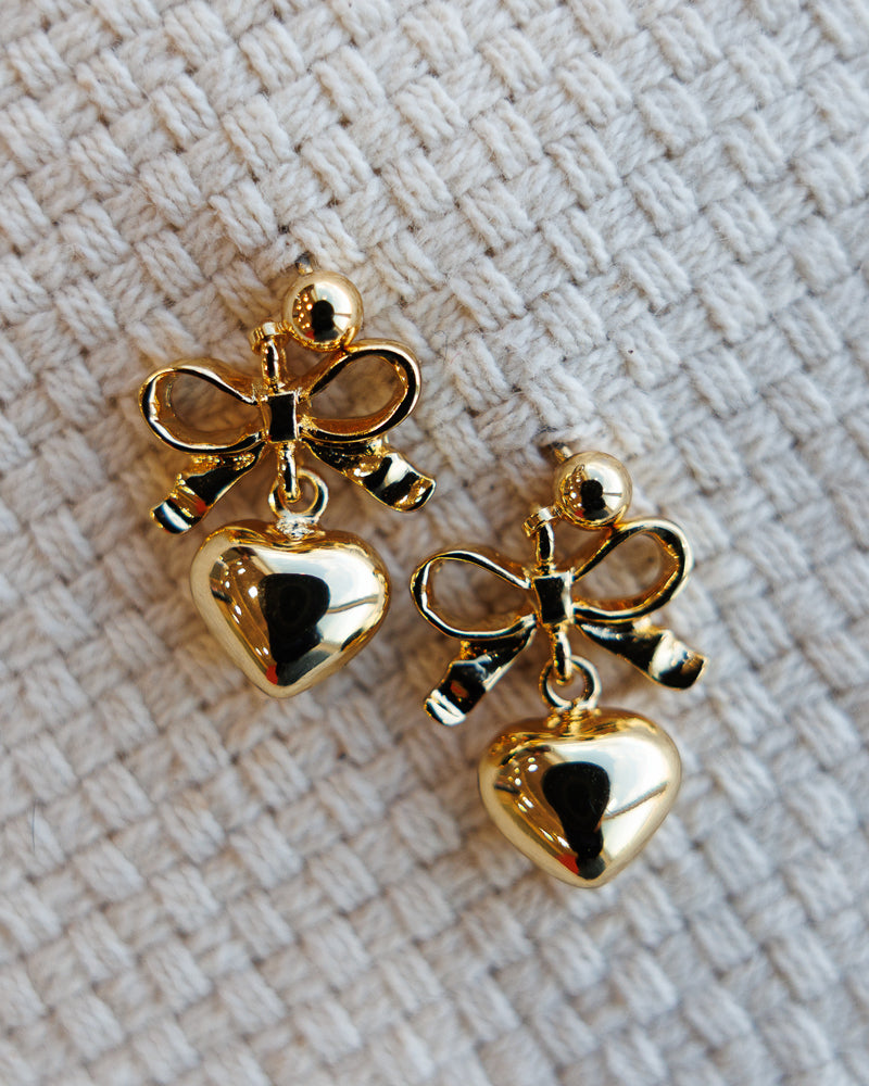 Gold heart-shaped earrings with bow details on a textured beige background