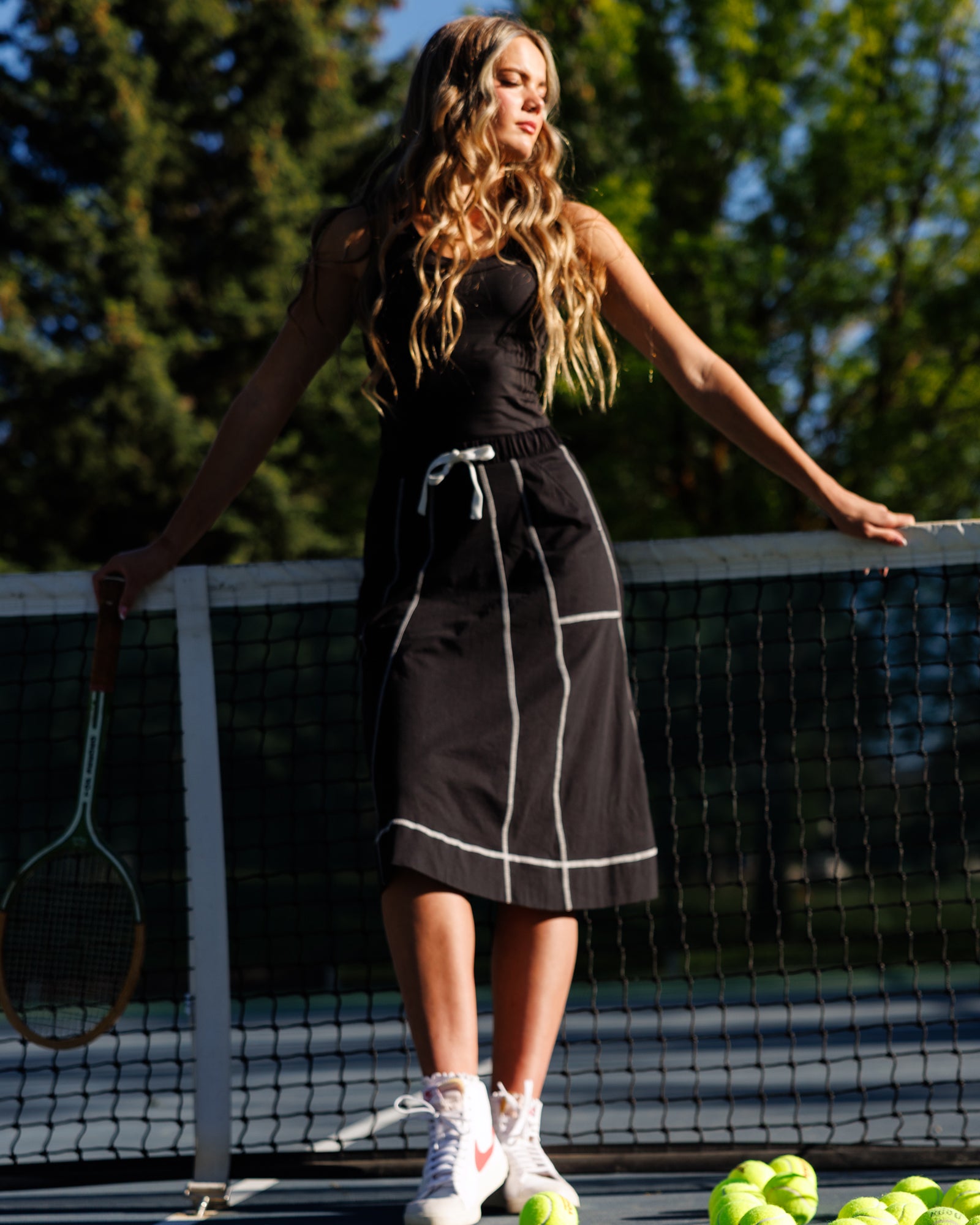 A woman stands on an outdoor tennis court holding a tennis racket, wearing a black sleeveless tennis dress with white piping details and white sneakers. Tennis balls are visible on the court floor, and trees are in the background.