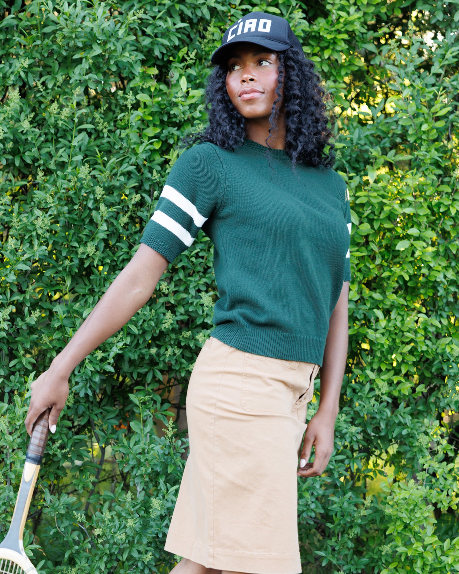 A woman wearing a dark green short-sleeve knit sweater with white stripes on the sleeves, a beige skirt, and a black baseball cap with 'CIRO' on it, standing outdoors in front of a green hedge and holding a tennis racket.