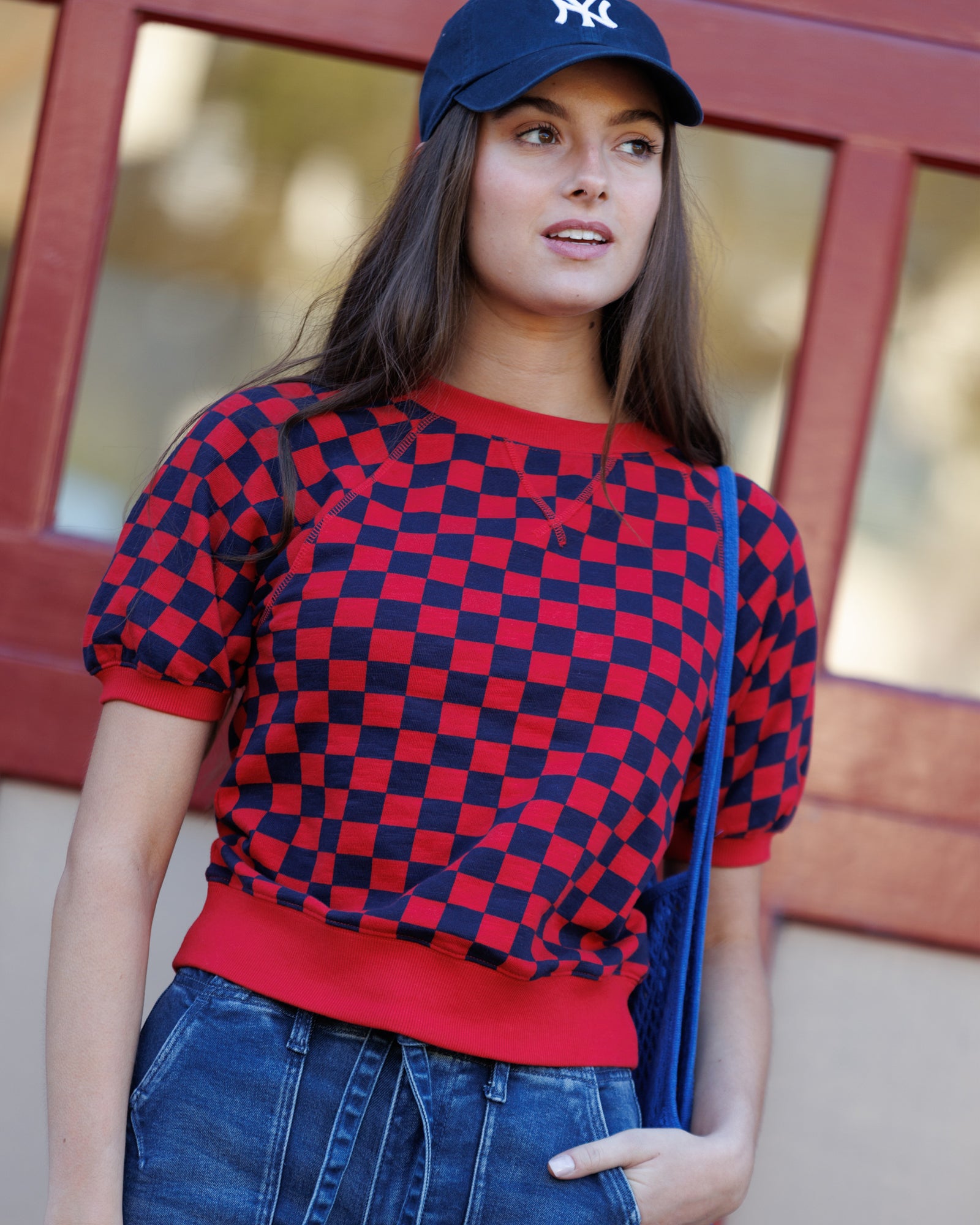 Woman wearing a red and blue checkered sweater with a blue cap and jeans, standing against a blurred background.