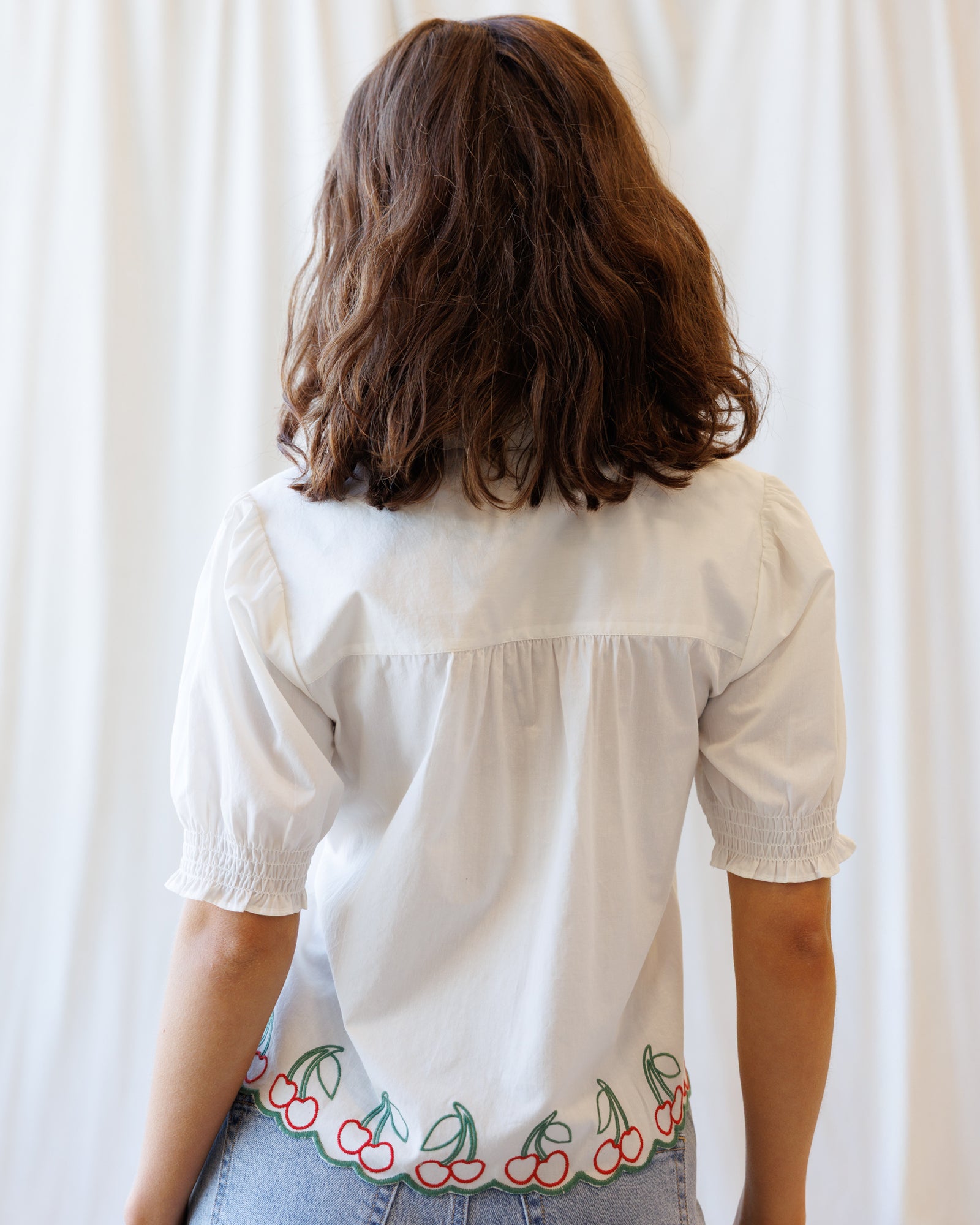 Person wearing a white blouse with cherry embroidery on a plain background