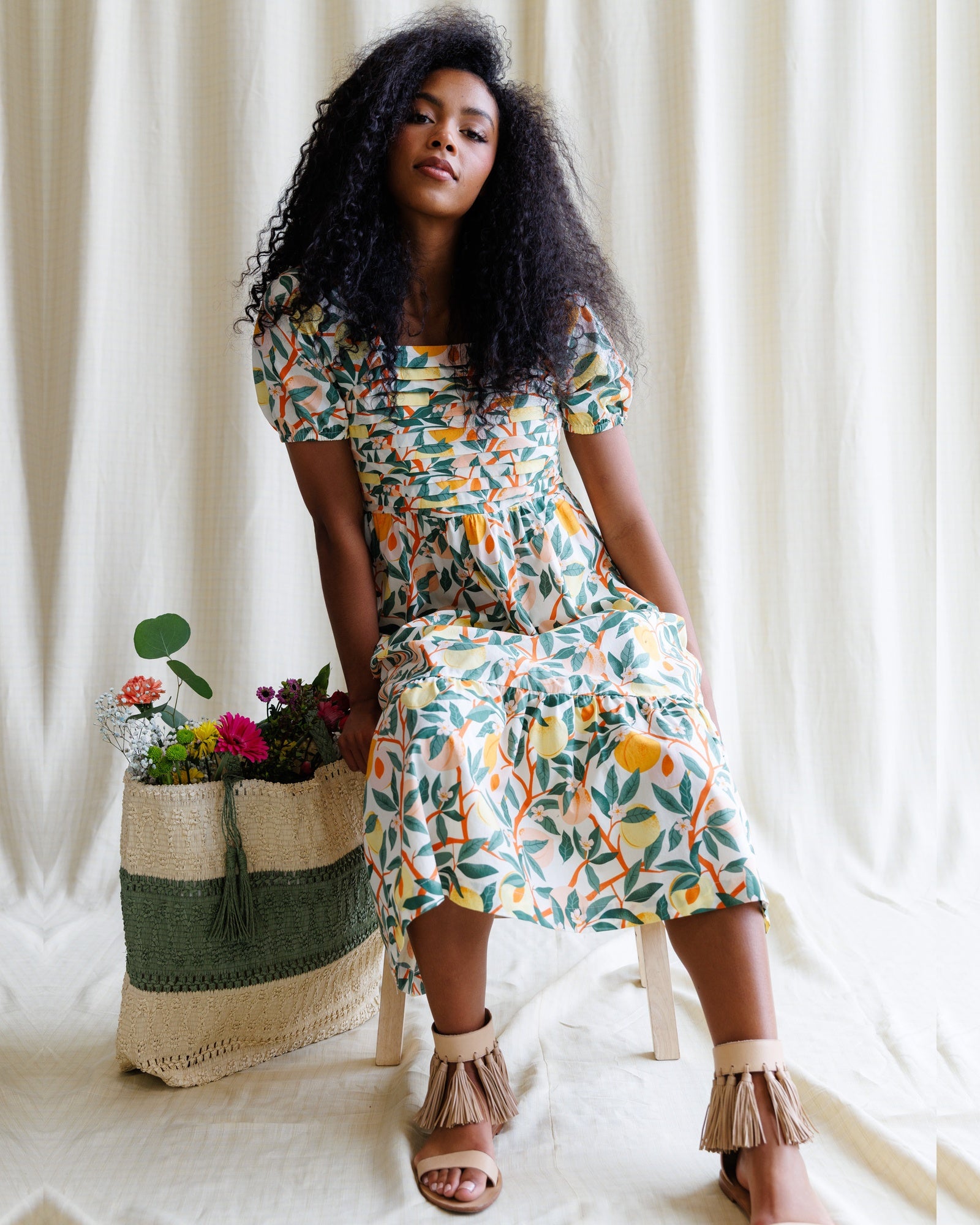 Woman in a colorful dress sitting next to a woven basket with flowers against a white curtain background