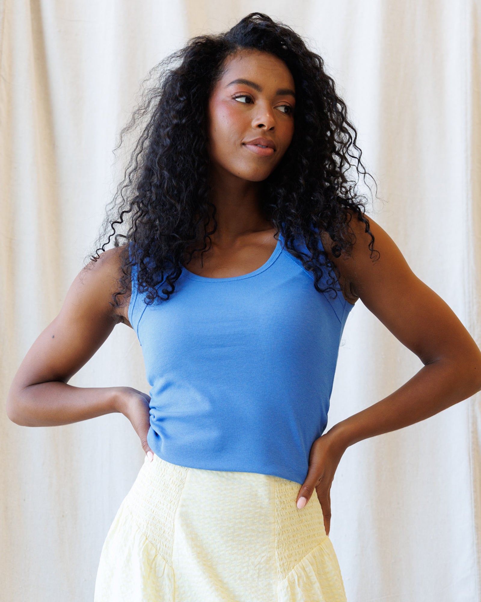 Woman wearing a blue tank top and yellow pants against a beige curtain background
