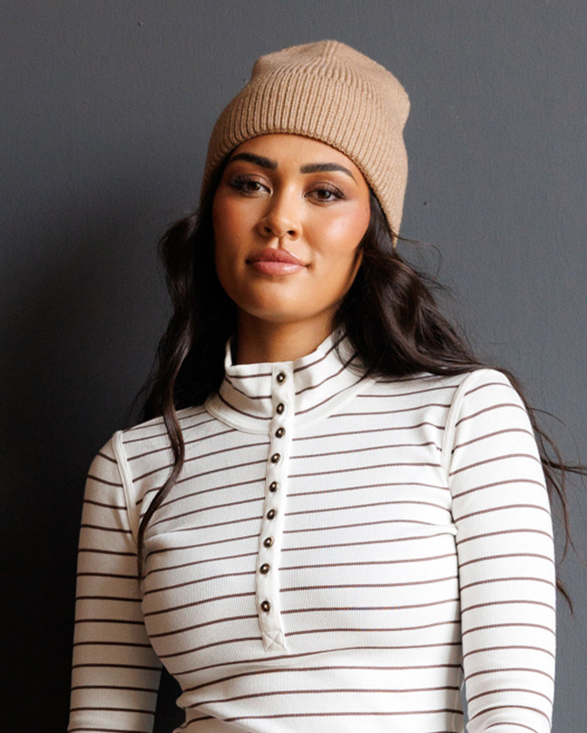 A woman wearing a beige ribbed knit beanie and a cream long-sleeve turtleneck top with thin brown horizontal stripes and a button placket, posed against a dark gray studio background.