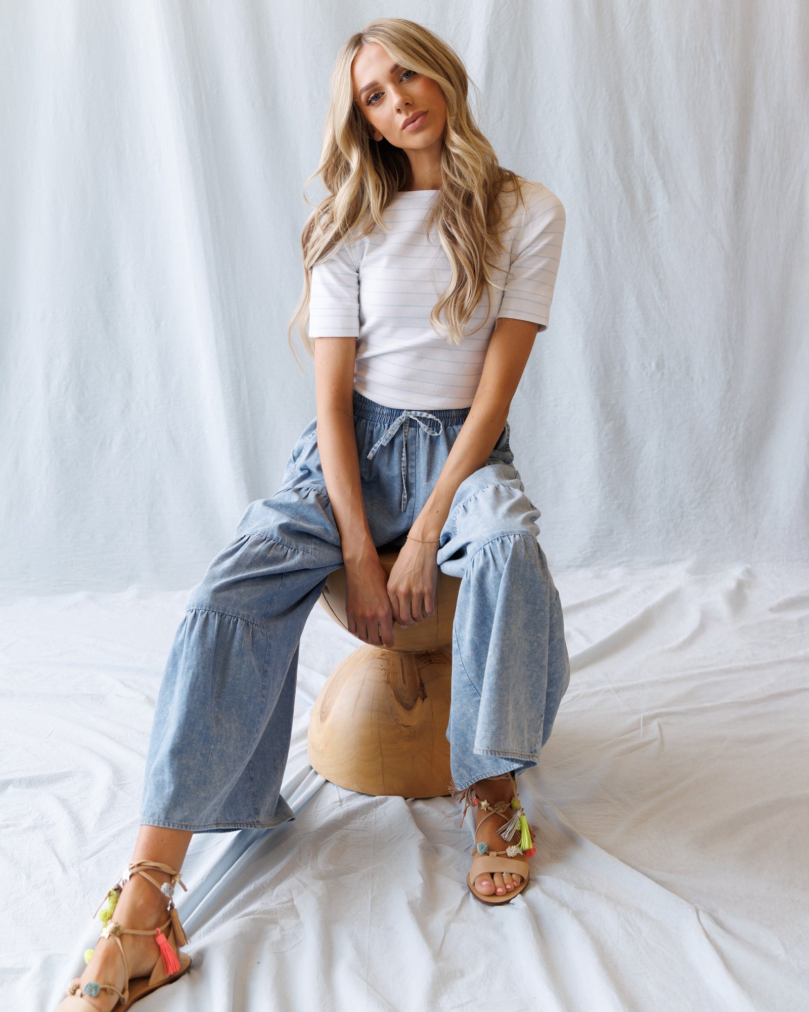 Woman sitting on a wooden stool wearing a white top and blue pants against a plain background