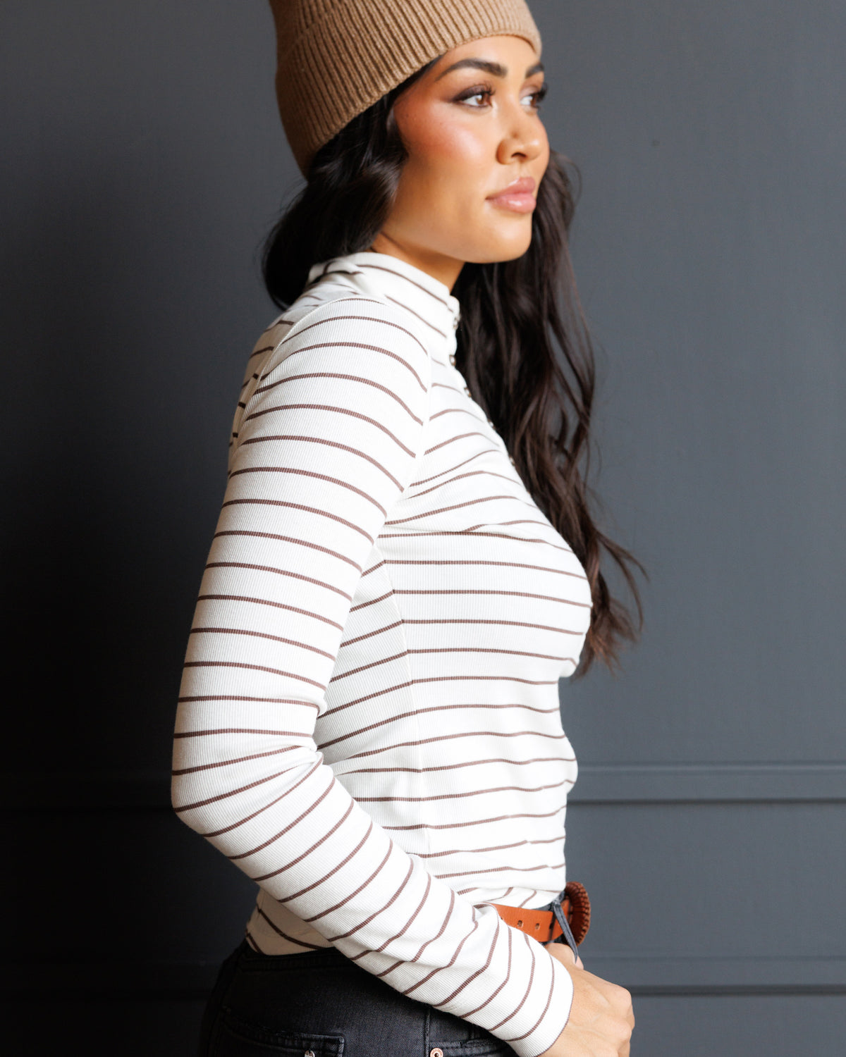 A woman in side profile wearing a fitted long-sleeve mock neck top with thin brown horizontal stripes on an off-white base, paired with a tan beanie and dark jeans, set against a dark gray paneled wall.