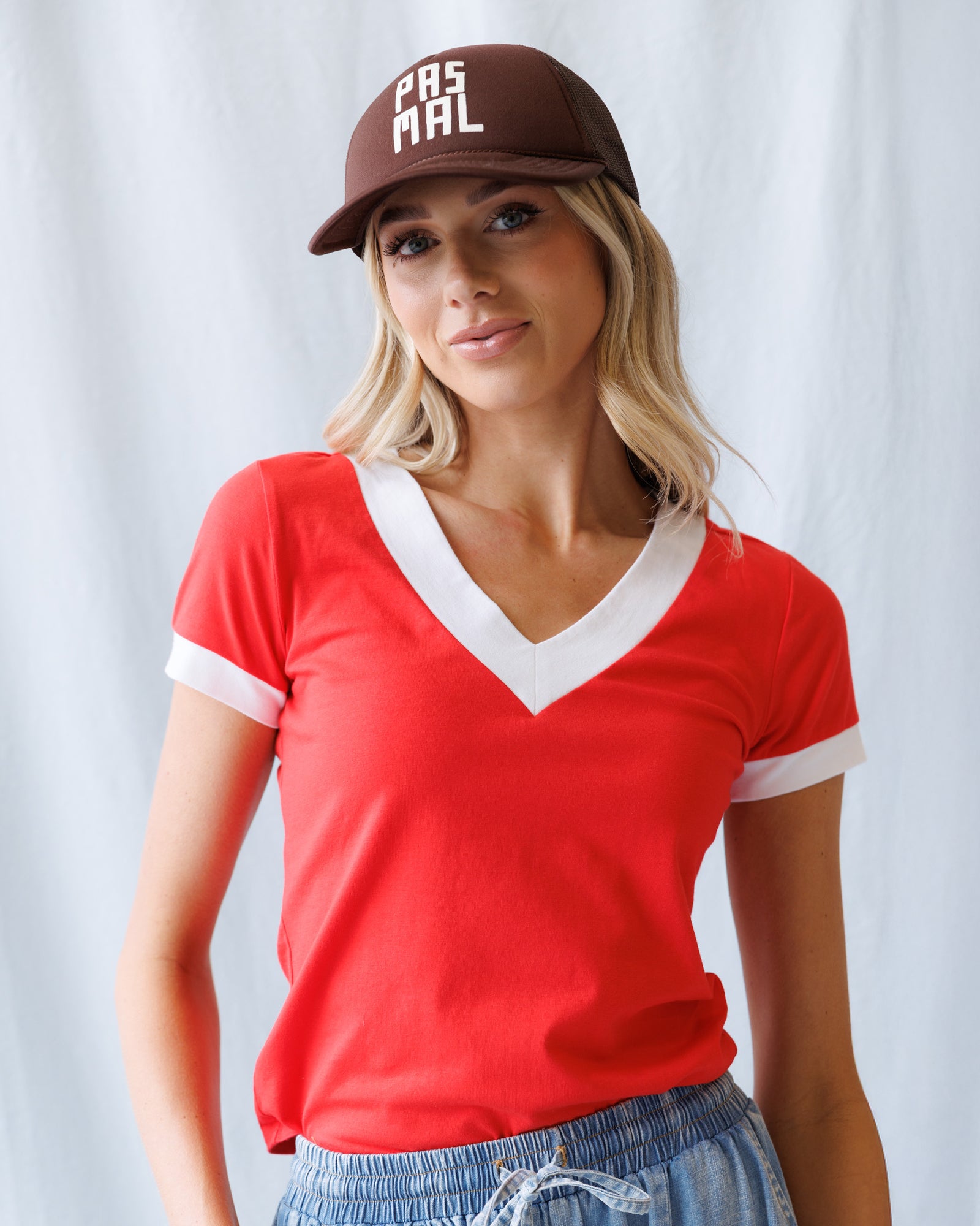 Woman wearing a red shirt with white trim and a brown cap with 'PAS MAL' logo against a white background