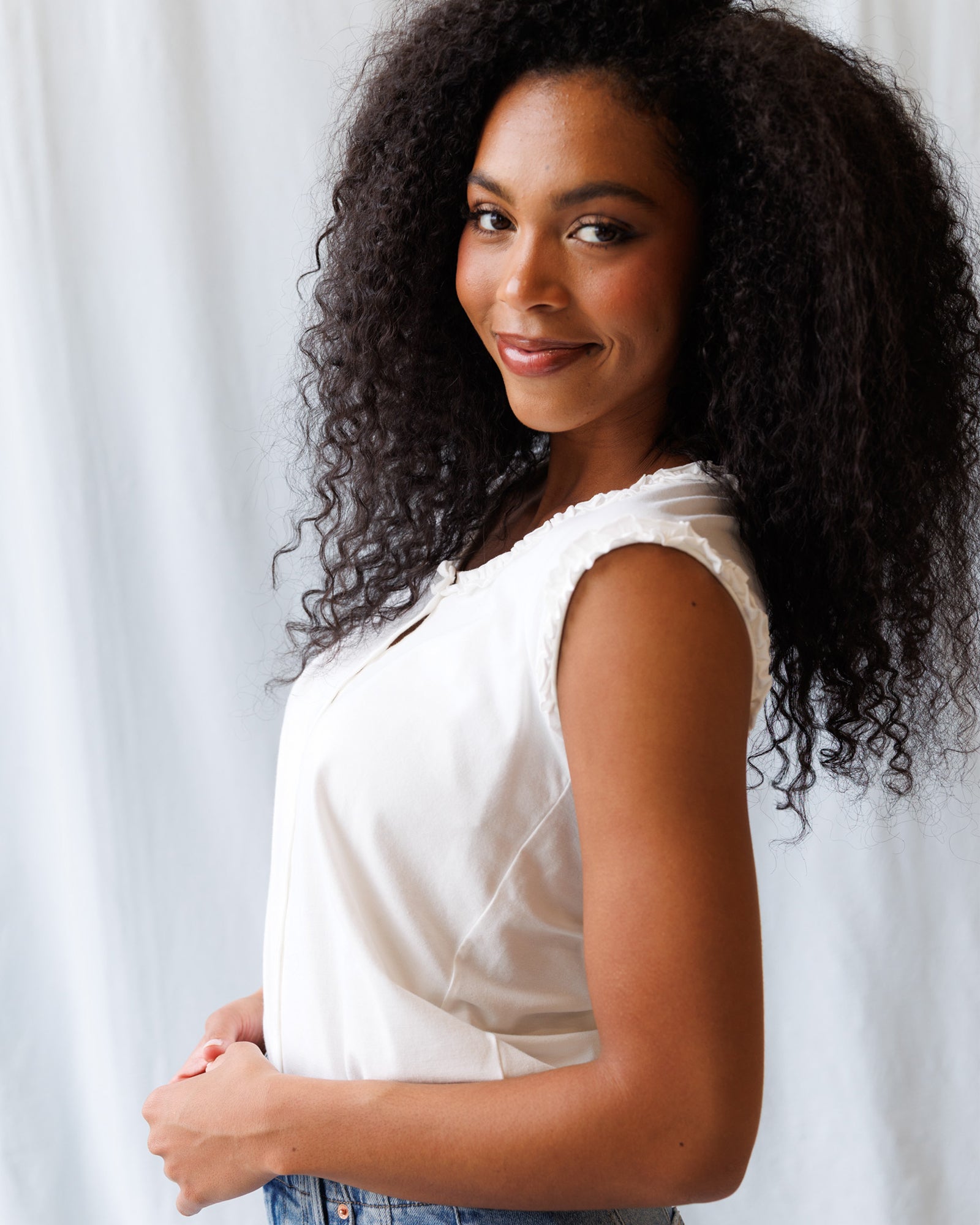 Woman with curly hair wearing a white sleeveless top against a light background