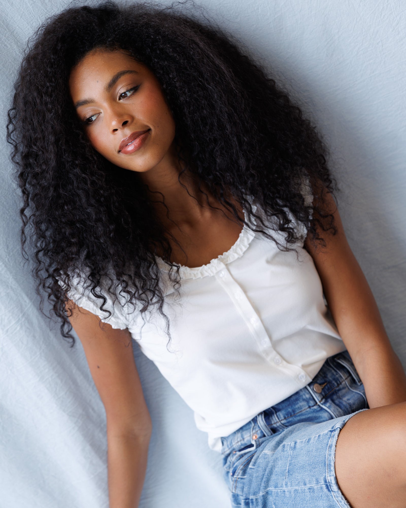 Woman with curly hair wearing a white top and denim shorts on a light background