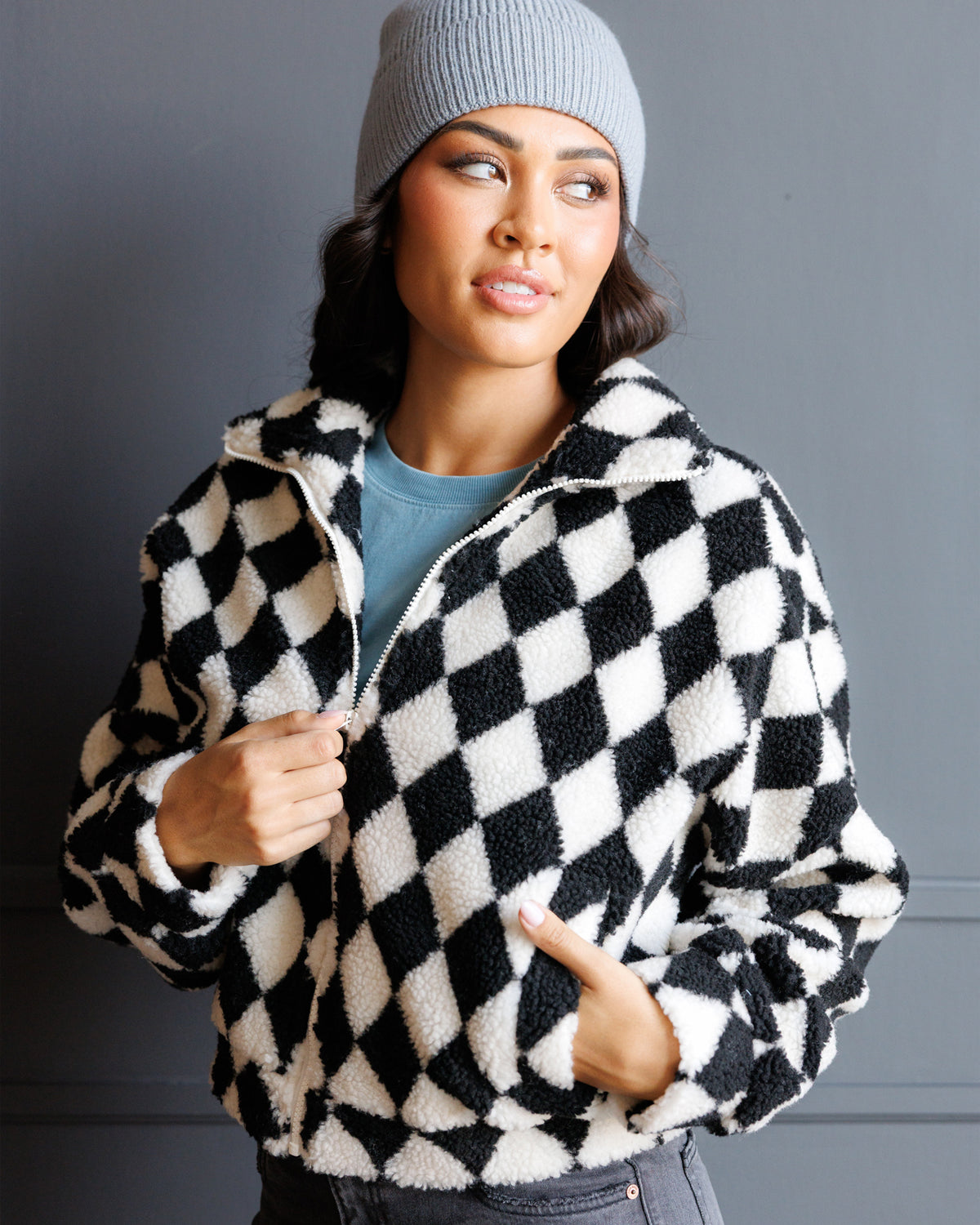 A woman wearing a black and white diamond-patterned sherpa fleece zip-up jacket, a light gray beanie, and a blue t-shirt, posing against a dark gray wall.