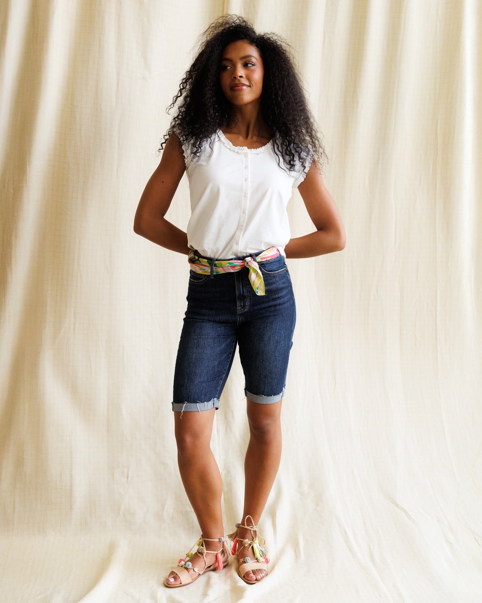 Woman wearing a white top and blue shorts with a colorful belt against a beige curtain background
