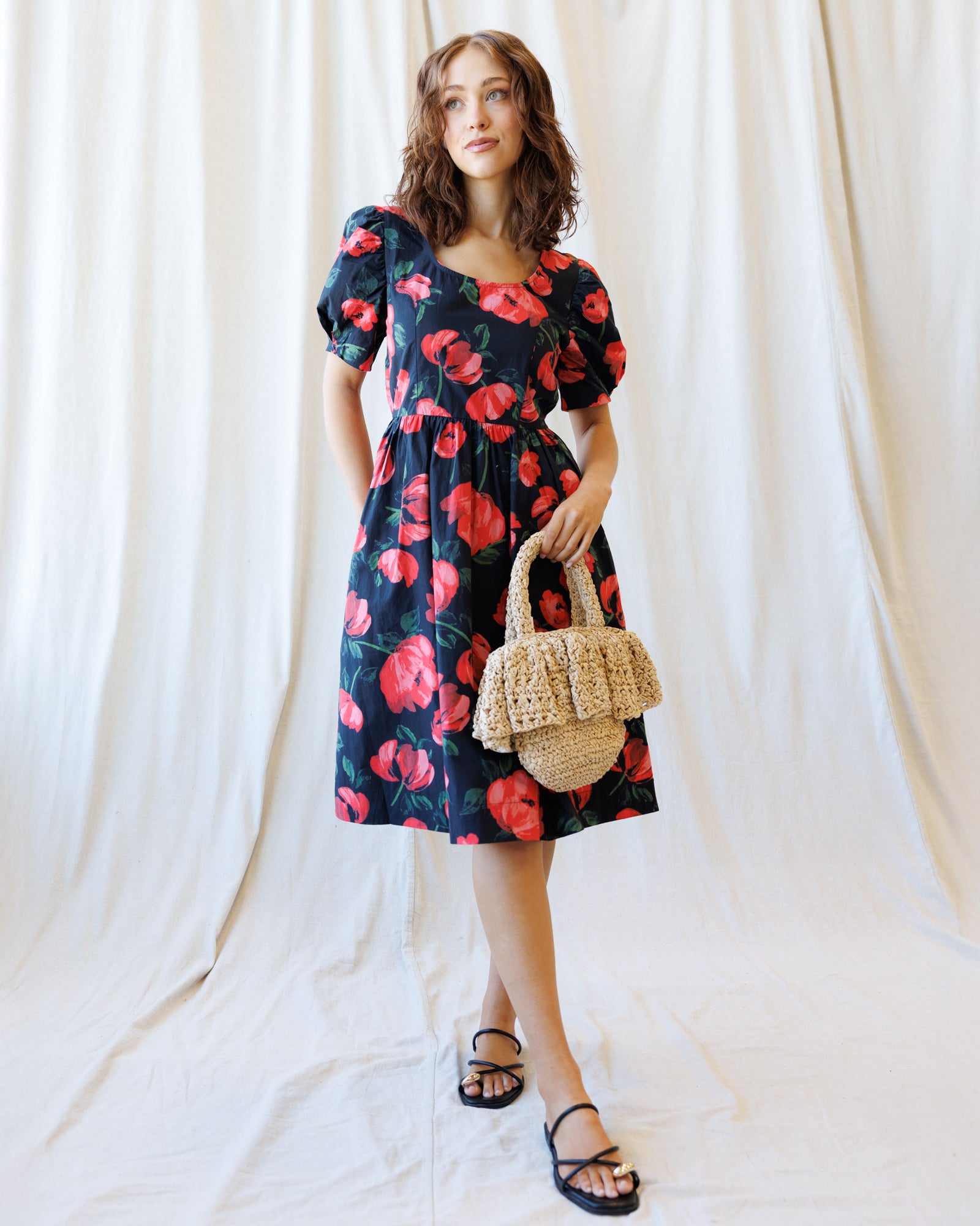 Woman wearing a floral dress holding a woven bag against a white curtain background
