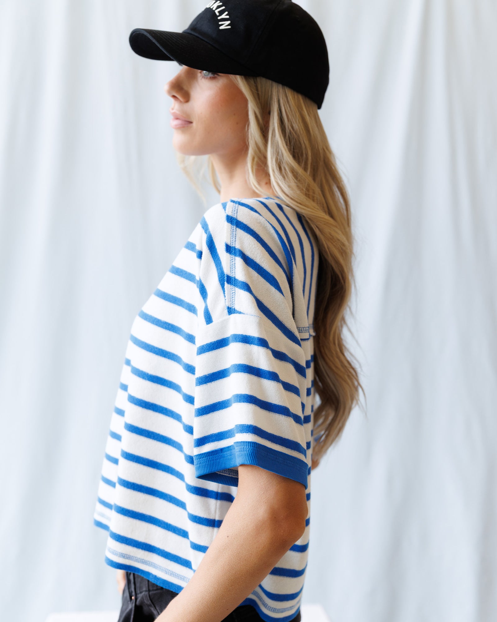 Person wearing a black cap and blue and white striped shirt against a white curtain background