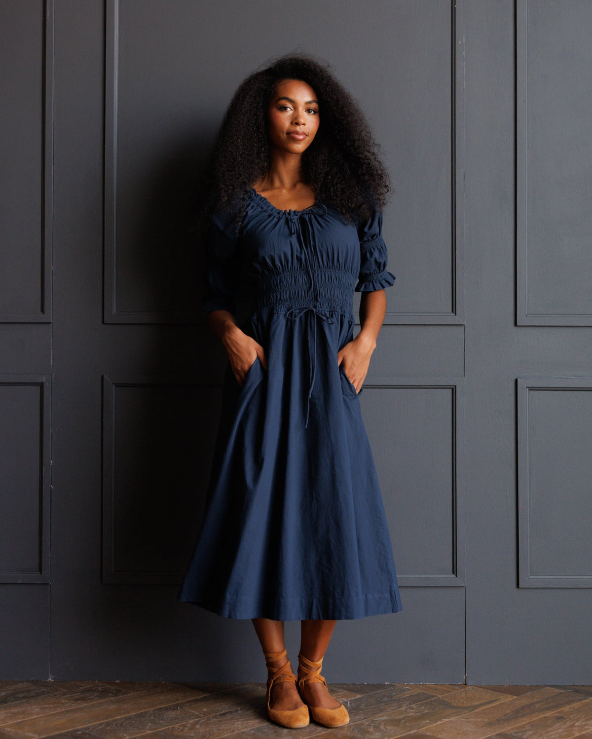 A woman with curly black hair stands against a dark gray paneled wall wearing a navy blue midi dress with ruffled sleeves and a gathered waist, paired with tan ankle boots.