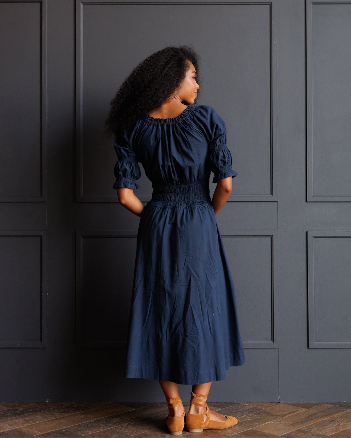 A woman stands with her back to the camera wearing a navy blue midi dress with puff sleeves and tan flat shoes, posing in front of a dark gray paneled wall.
