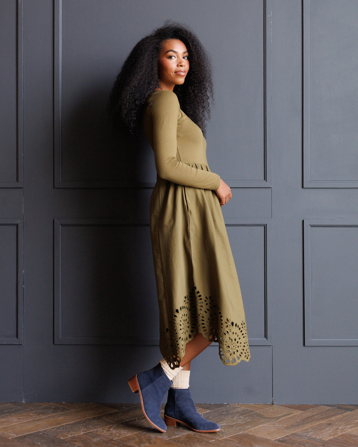 A woman with curly black hair stands in profile against a dark gray paneled wall, wearing a long-sleeve olive green midi dress with a scalloped eyelet hem and blue ankle boots.