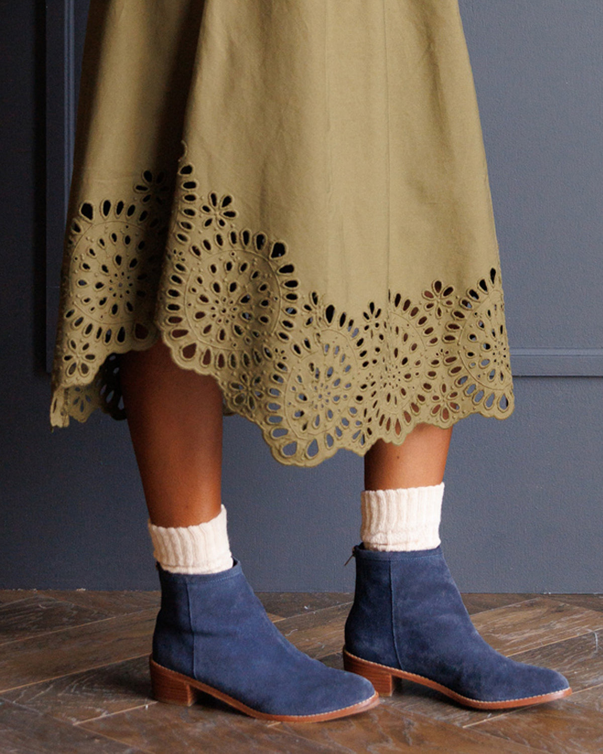 A person wearing a muted olive green midi skirt with a scalloped eyelet lace hem, paired with cream ribbed socks and navy blue suede ankle boots with brown leather soles, standing on a wooden floor against a dark gray wall.